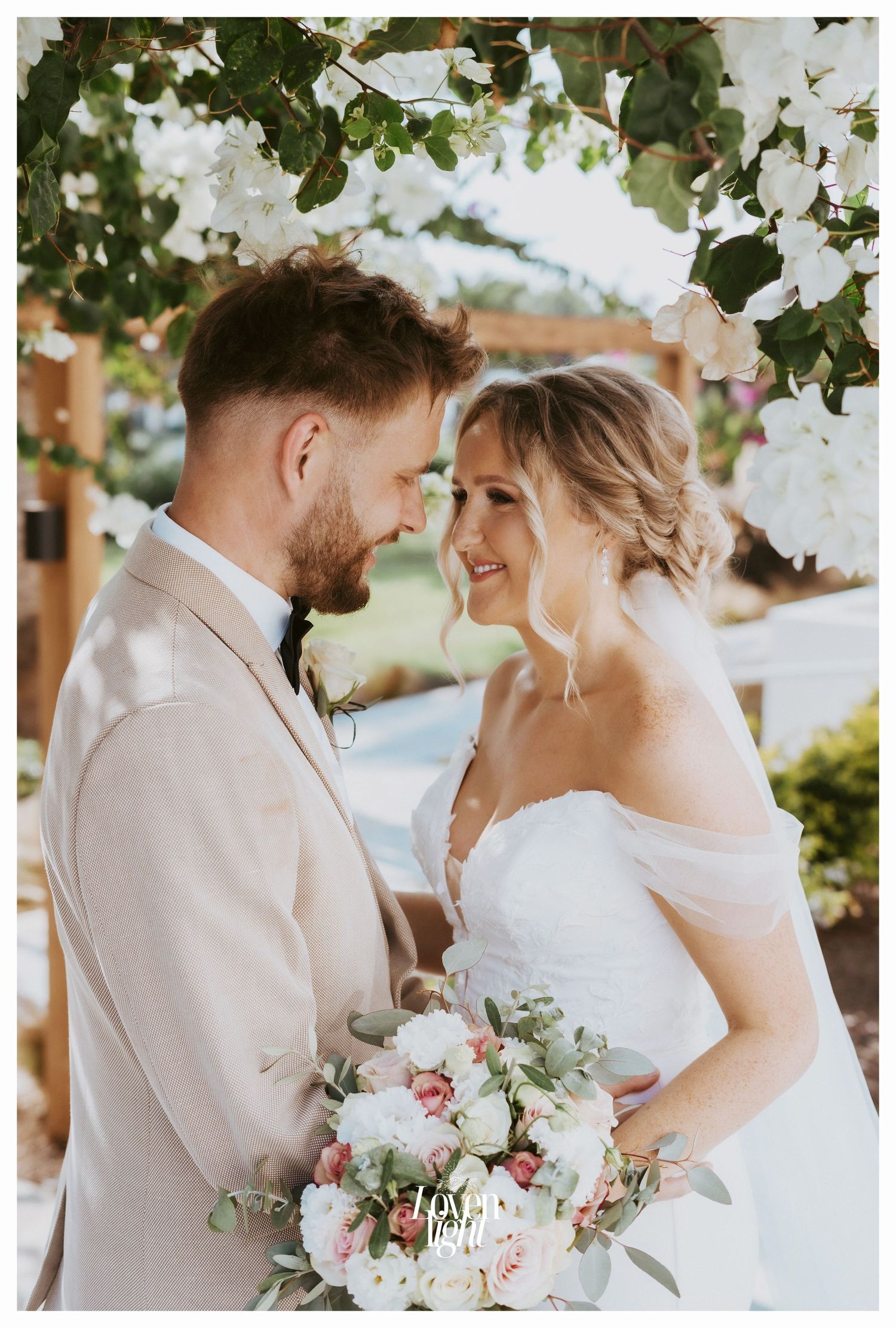 Close-up wedding photo of a couple smiling at each other beneath white flowering branches.
