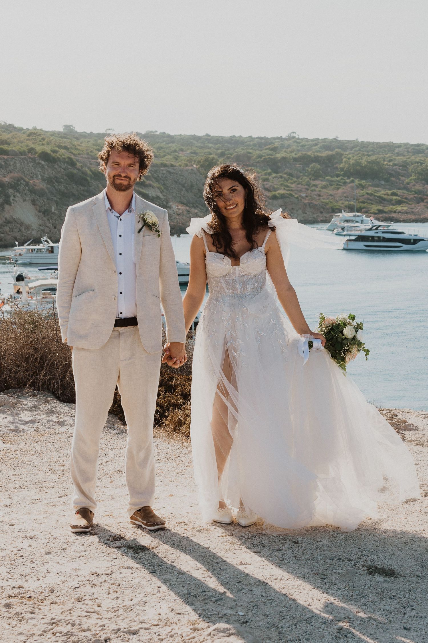 A newlywed couple in elegant white attire poses together on a cliff overlooking boats in a harbor.
