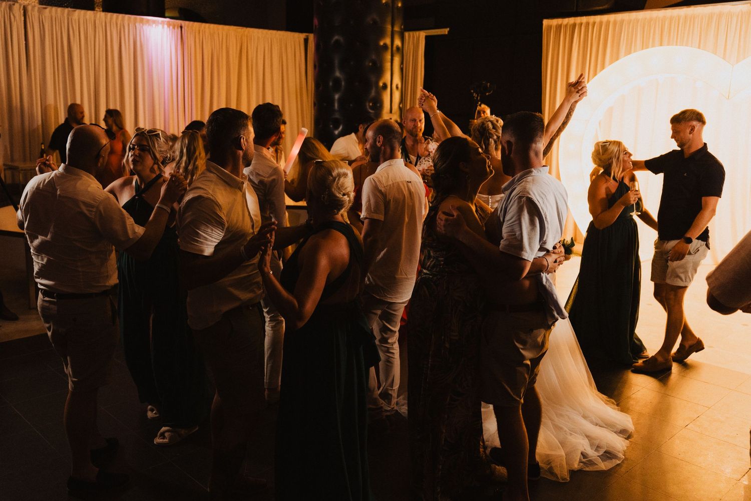 Wedding guests gather on the dance floor during an evening reception celebration.