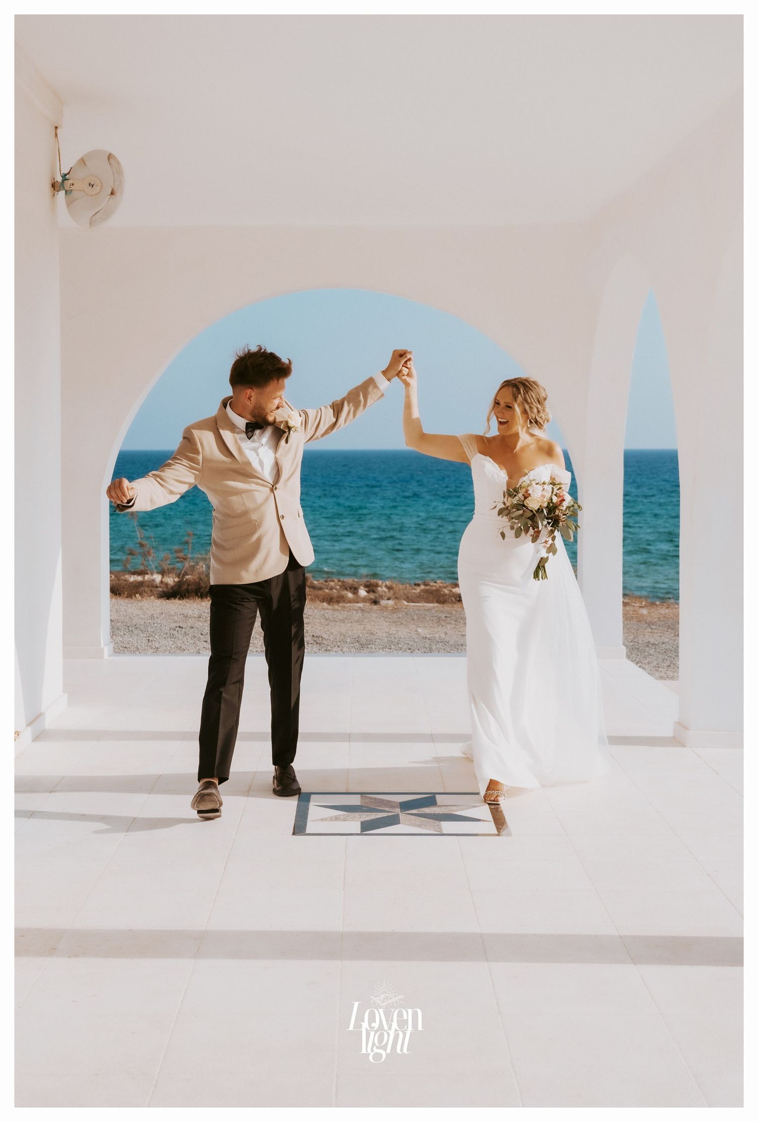 Joyful wedding couple dancing under white archway with ocean vista behind them.