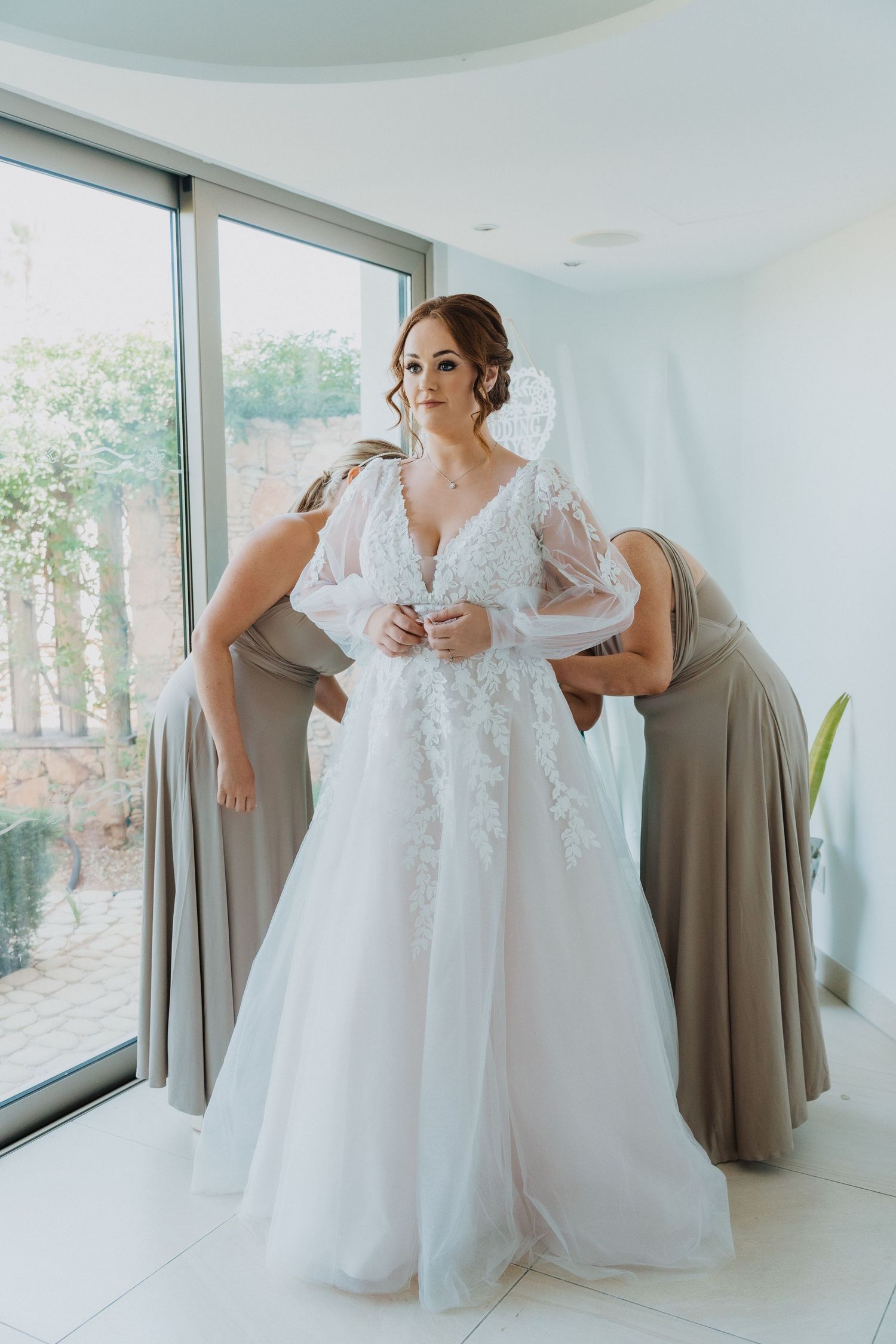 Wedding dress preparation moment in a bright white room with large windows.