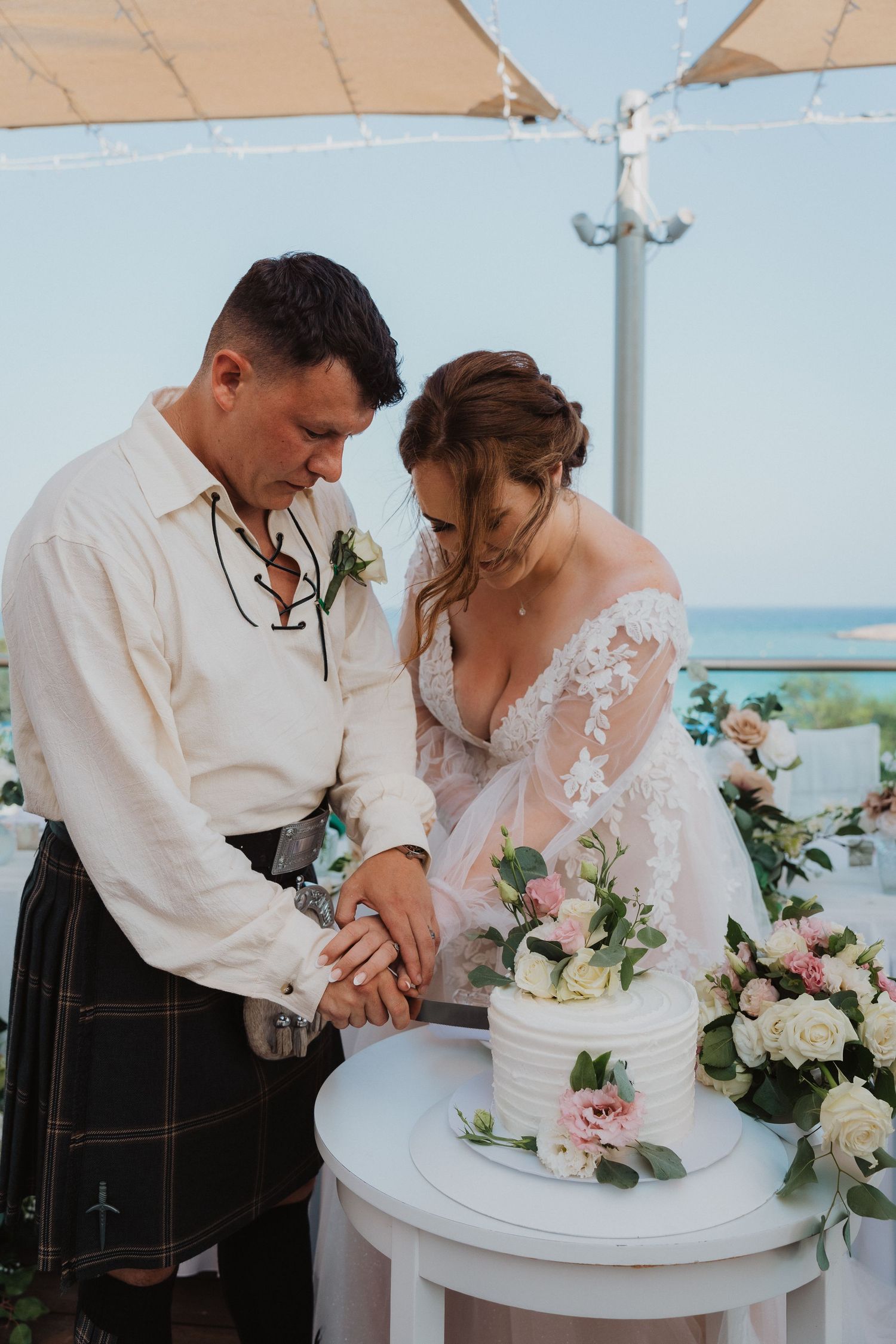 Wedding couple cutting their white floral-decorated cake at seaside venue.