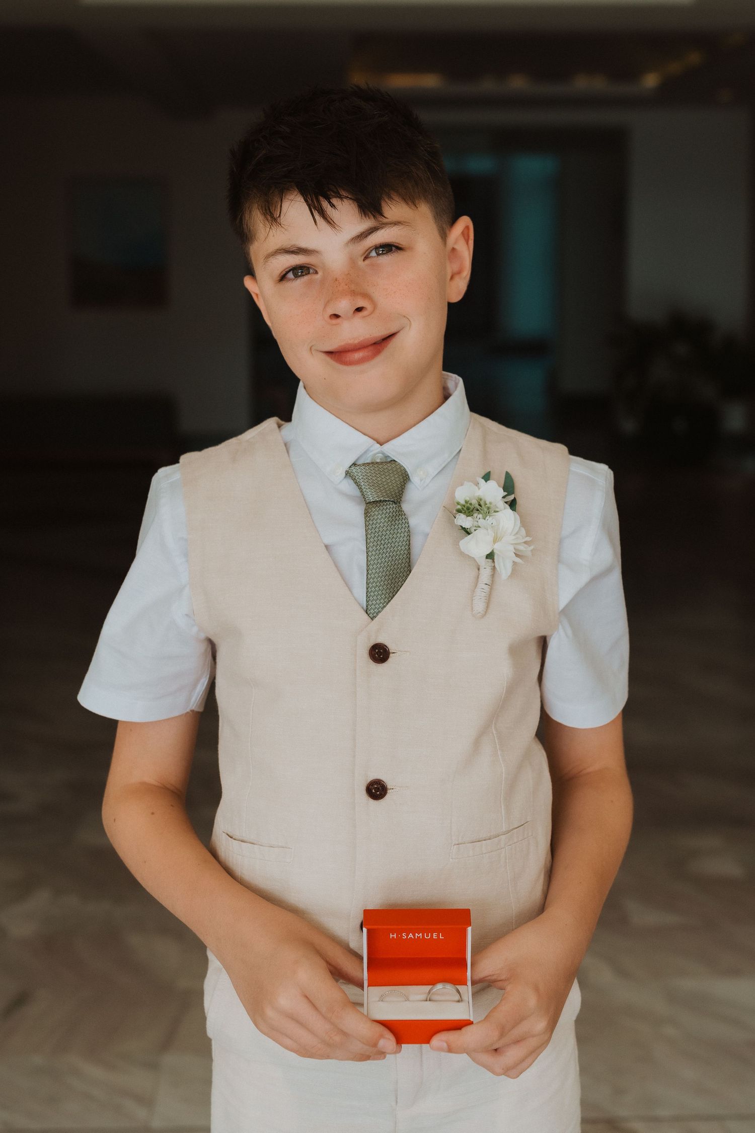Young wedding attendant in beige vest and sage tie holds red ring box while wearing a white boutonniere.
