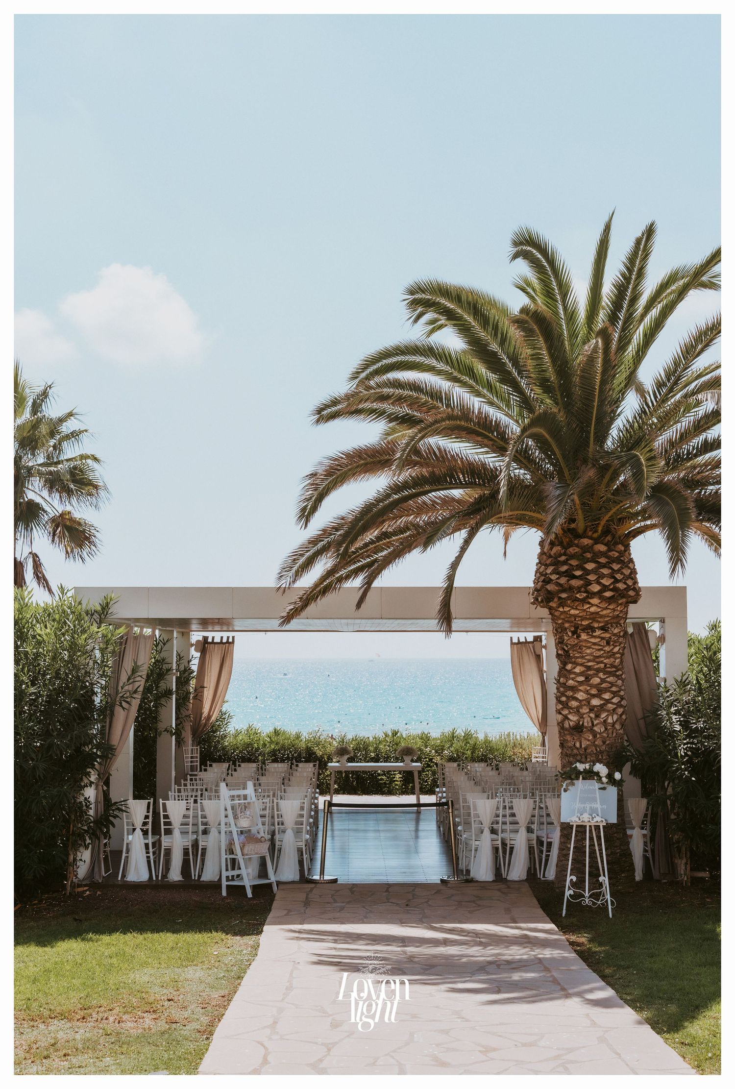 Wedding ceremony setup with white chairs and fabric under palm trees overlooking the sea.