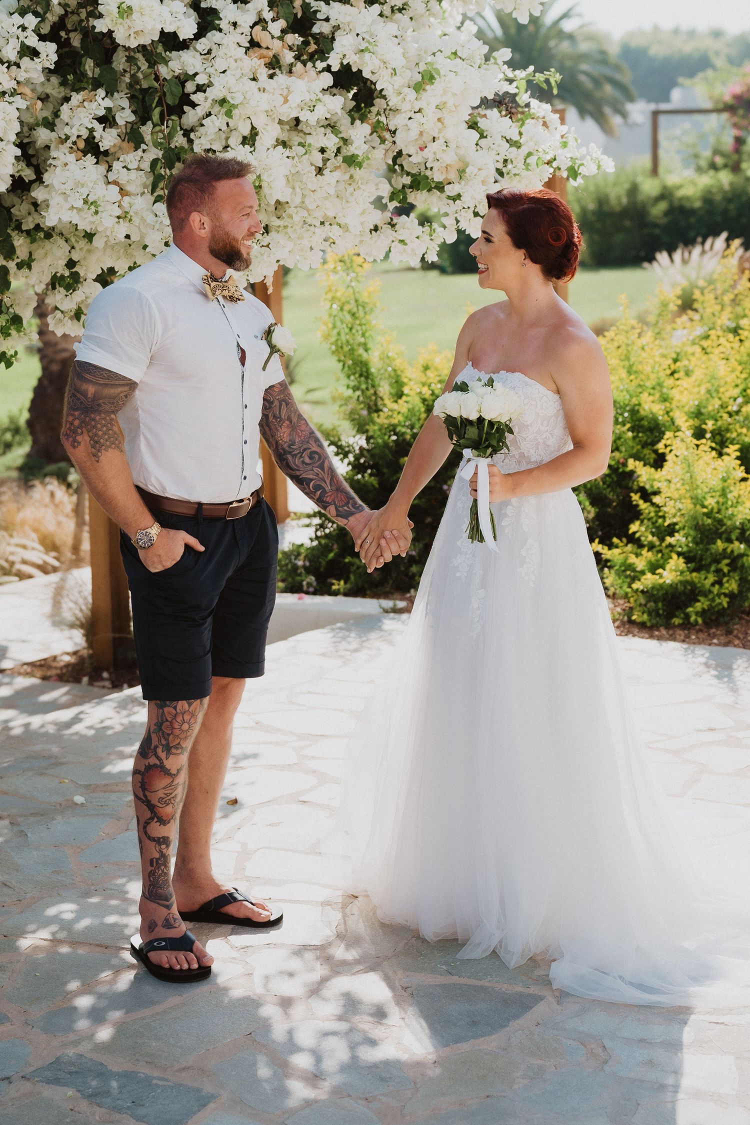 A couple shares a joyful moment under white floral blooms on their wedding day in a tropical garden setting.