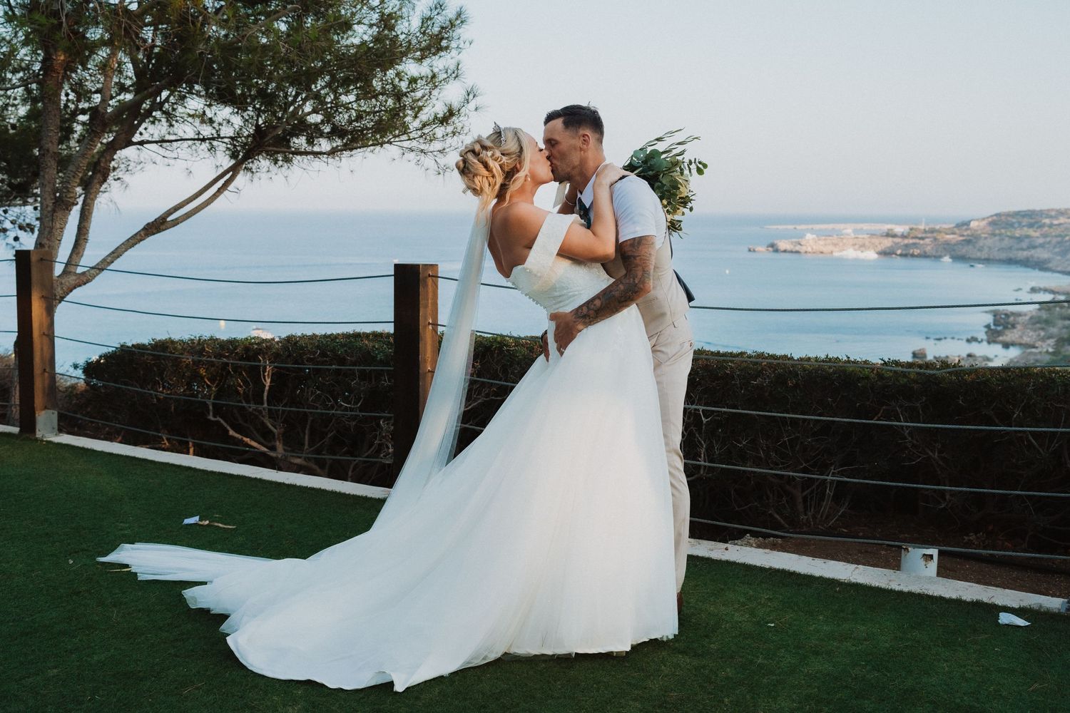 Wedding couple embraces on lawn overlooking coastal waters during sunset.