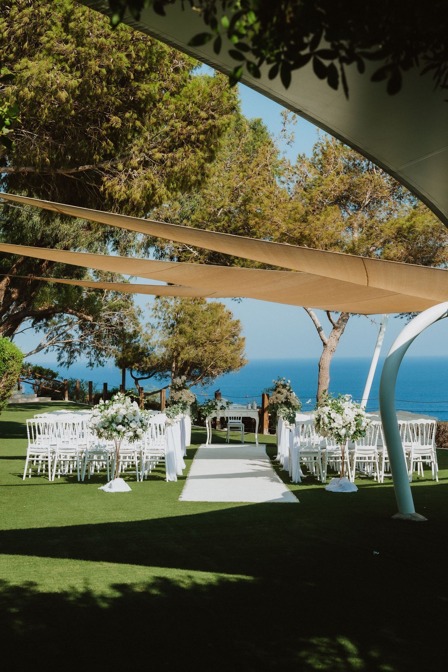 Outdoor wedding ceremony setup with white chairs and ocean views under elegant shade sails.