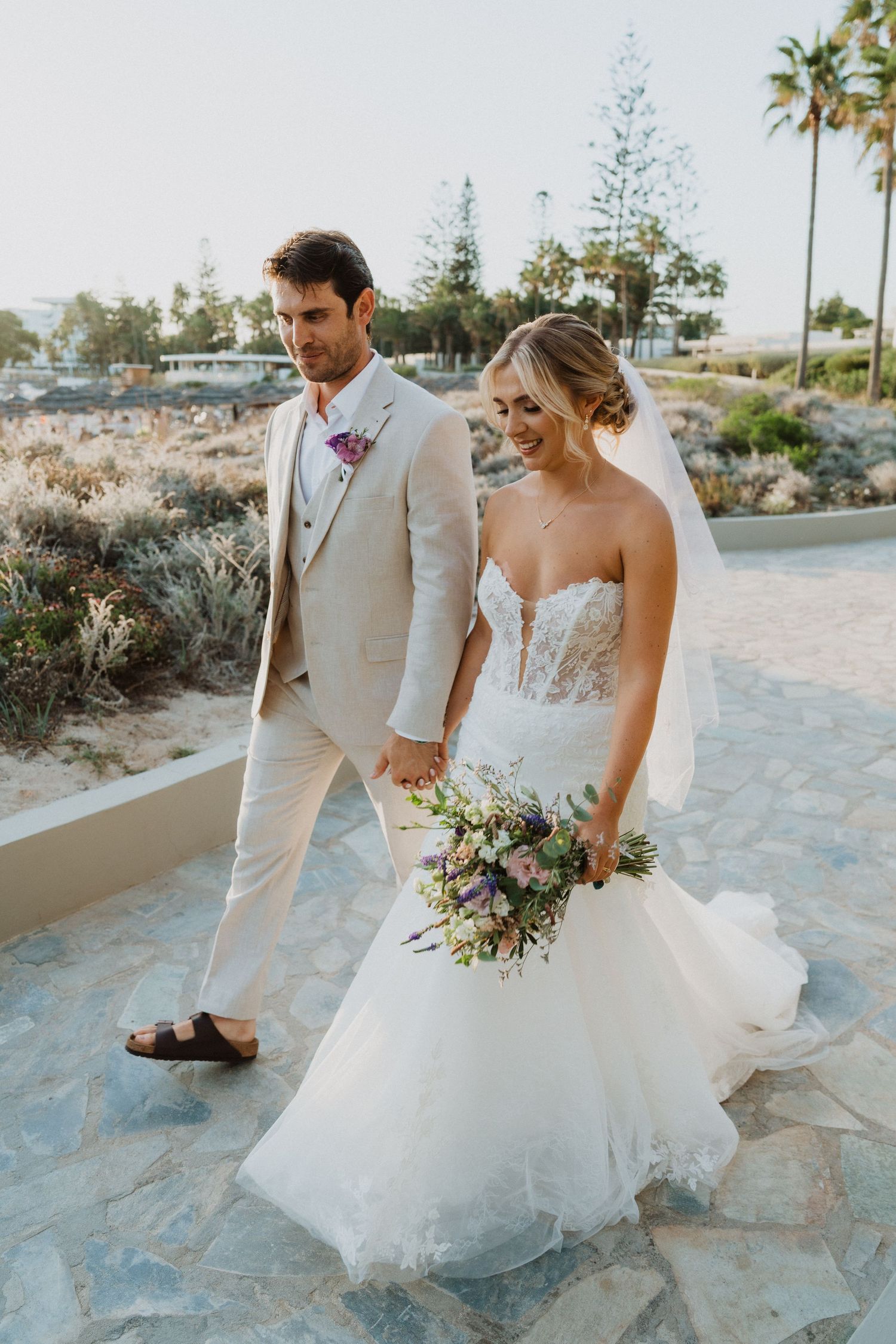 Wedding couple walking on stone pathway with bride holding bouquet and groom in light suit.