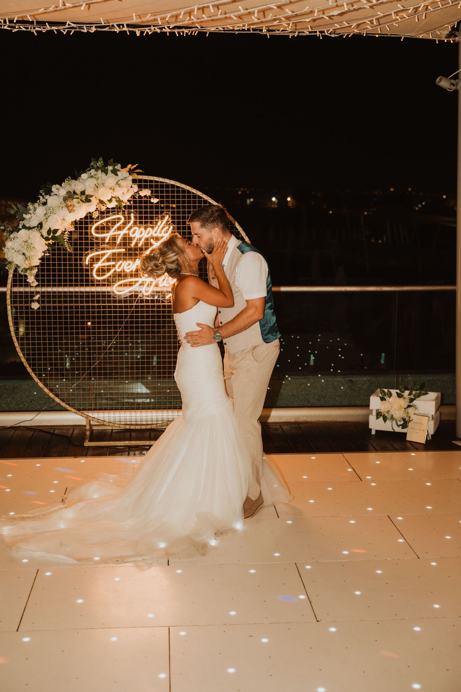 Bride and groom share a romantic first dance kiss under a glowing Happily Ever After neon sign at their wedding reception.