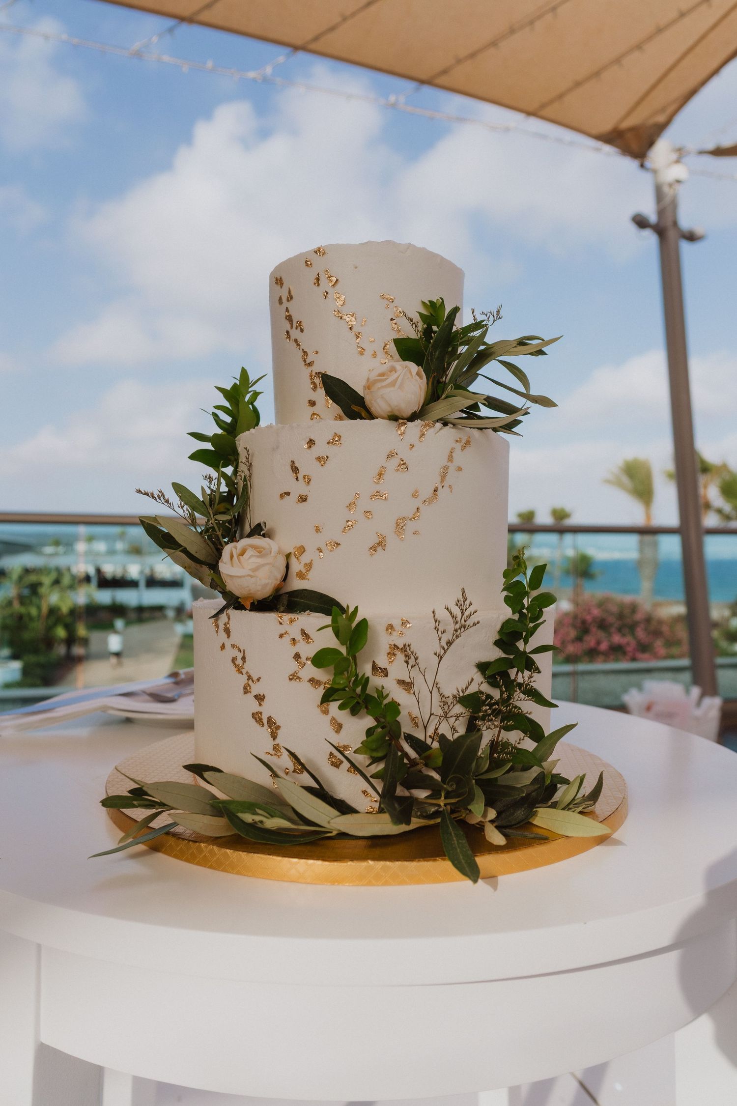 Elegant white wedding cake with gold accents and green foliage displayed outdoors under a covered patio.