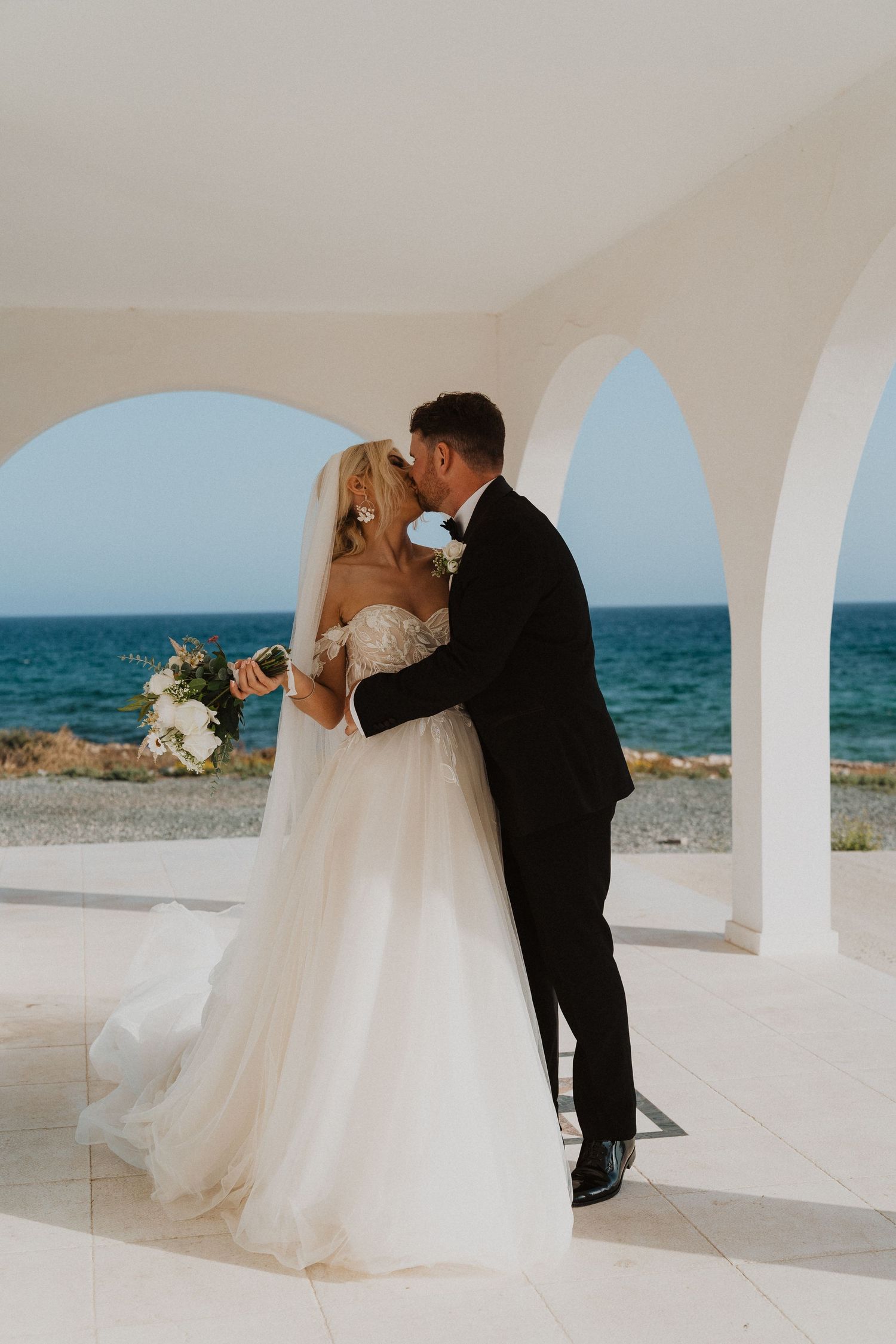 Wedding couple shares romantic moment under Mediterranean arches with ocean backdrop.