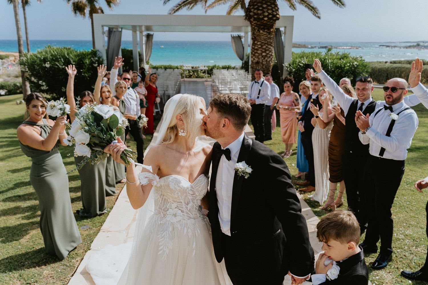 Newlyweds celebrate with guests during an outdoor beachfront wedding ceremony surrounded by palm trees.
