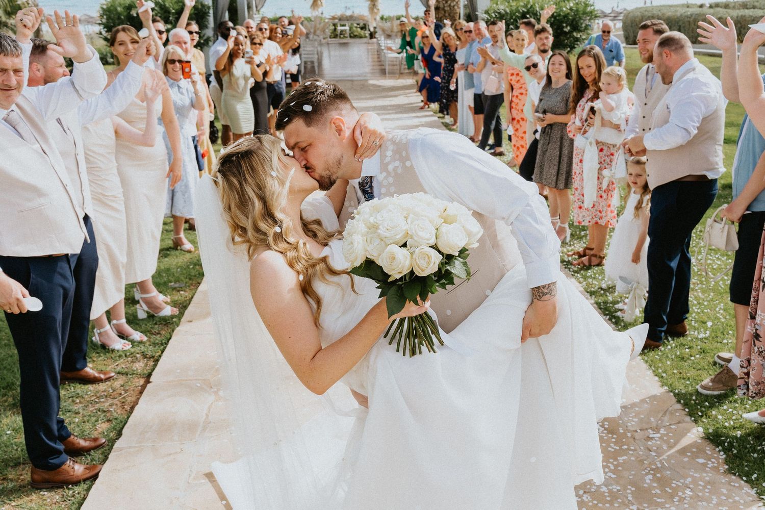 A romantic wedding day kiss with guests lined up along a pathway, while the bride holds a white bouquet of roses.