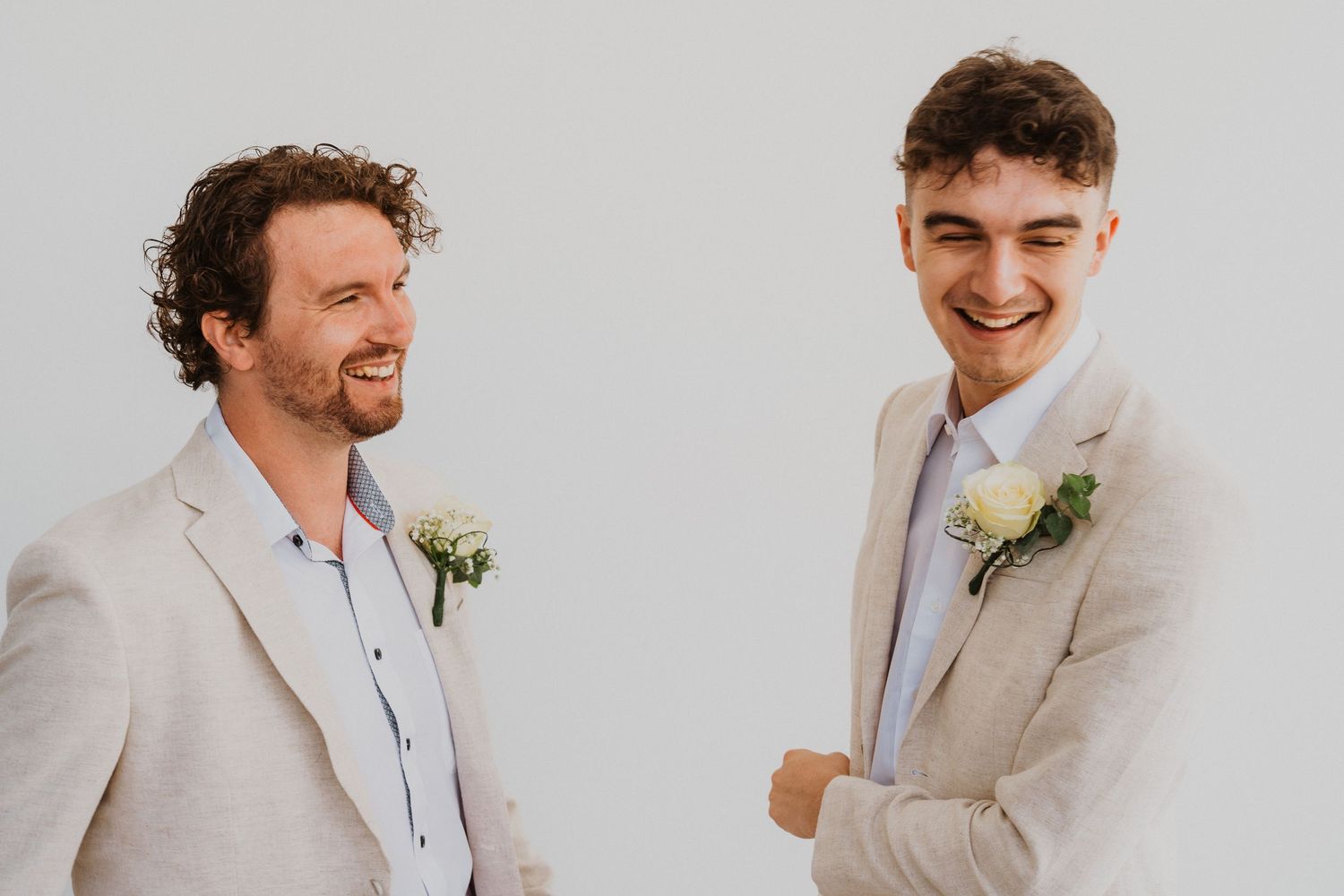 Two groomsmen in light colored suits share a candid laughing moment during wedding preparations.