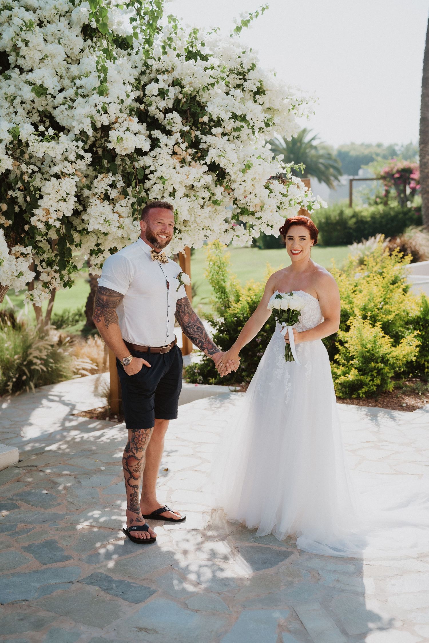 Casual beach wedding couple holding hands on stone patio with flowering arbor.
