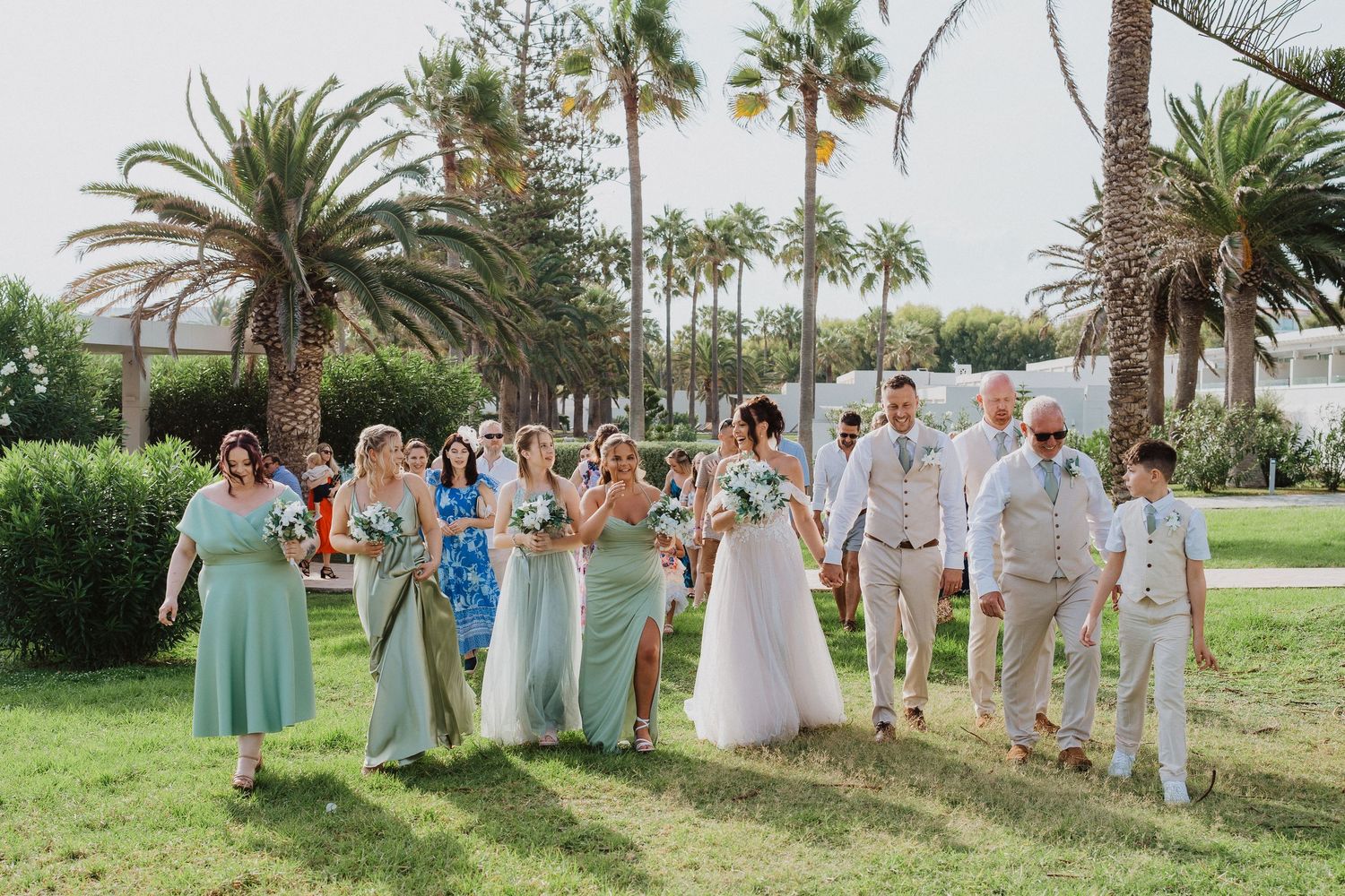 Wedding party poses on lush green lawn with palm trees and tropical landscaping in the background.
