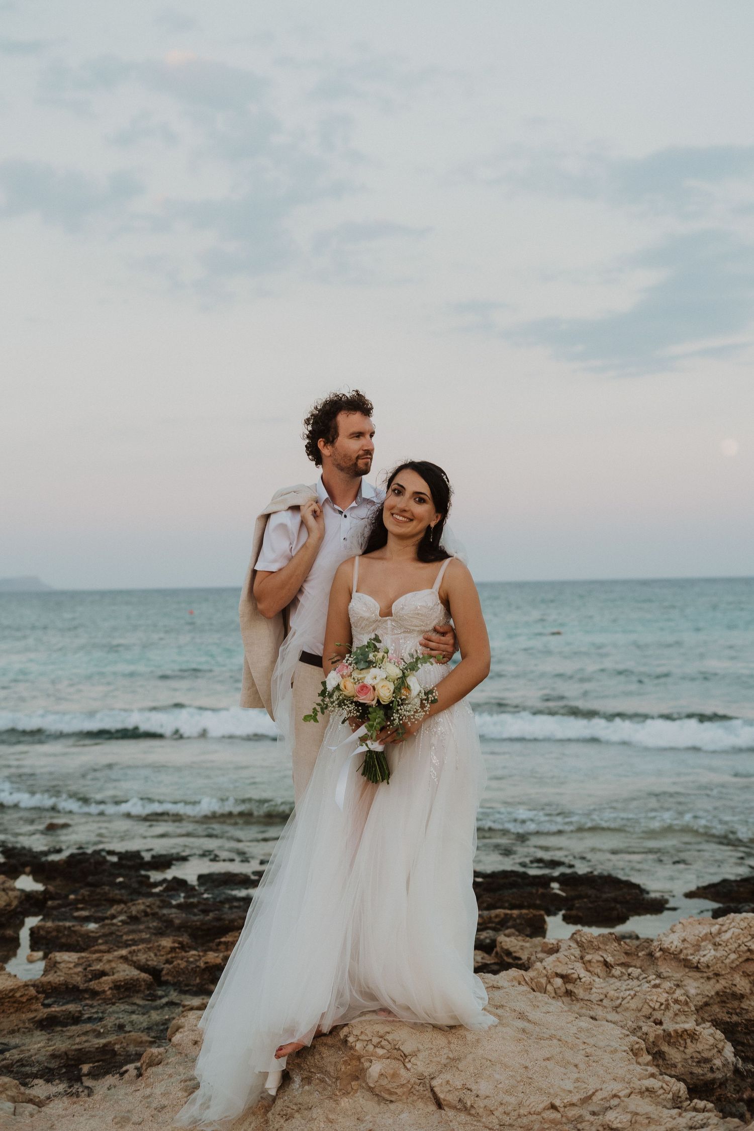 A joyful wedding portrait on rocky beach terrain with turquoise ocean waters in the background.
