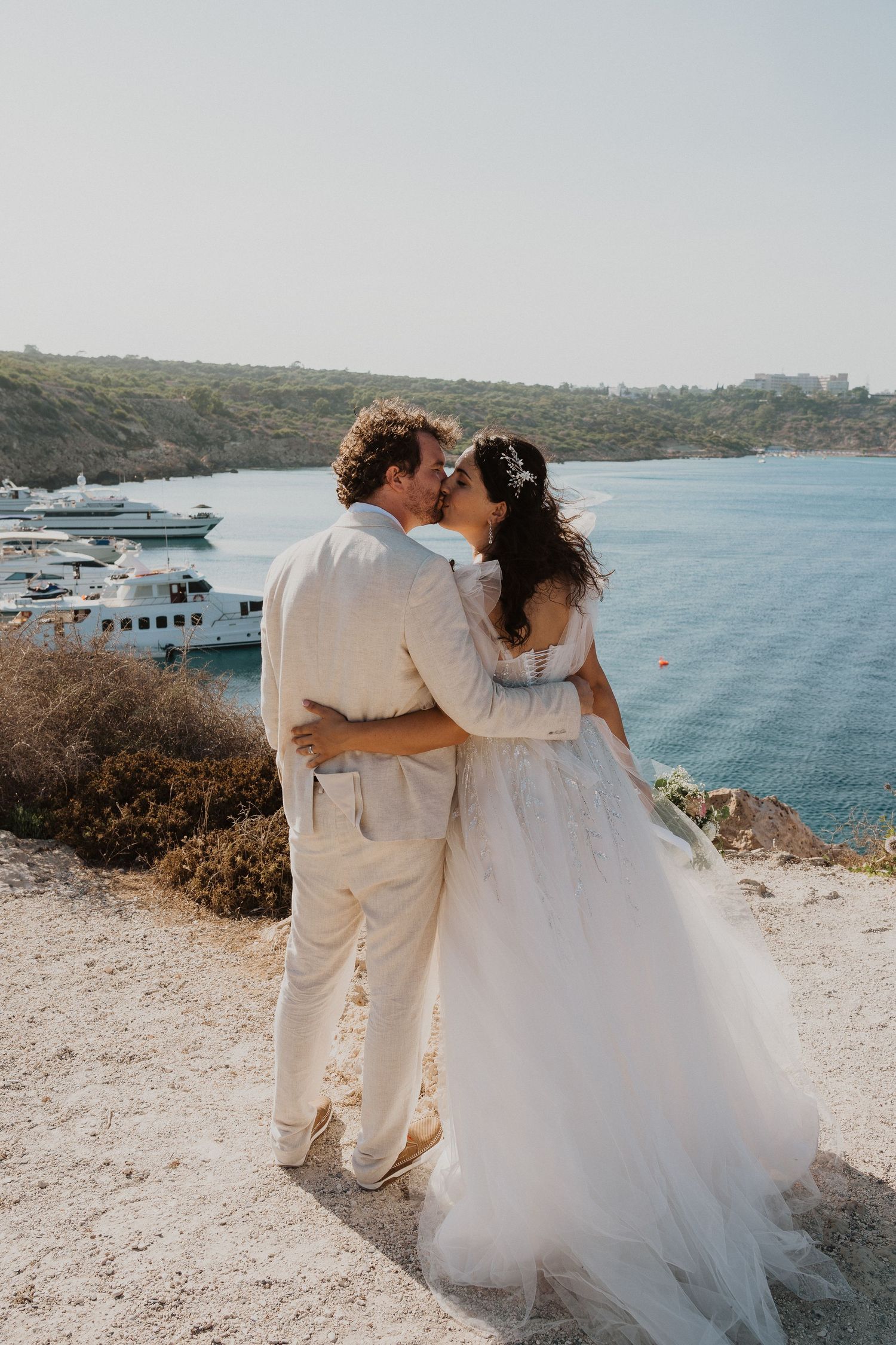A romantic wedding couple embraces while overlooking a scenic marina bay on a sunny coastal cliff.
