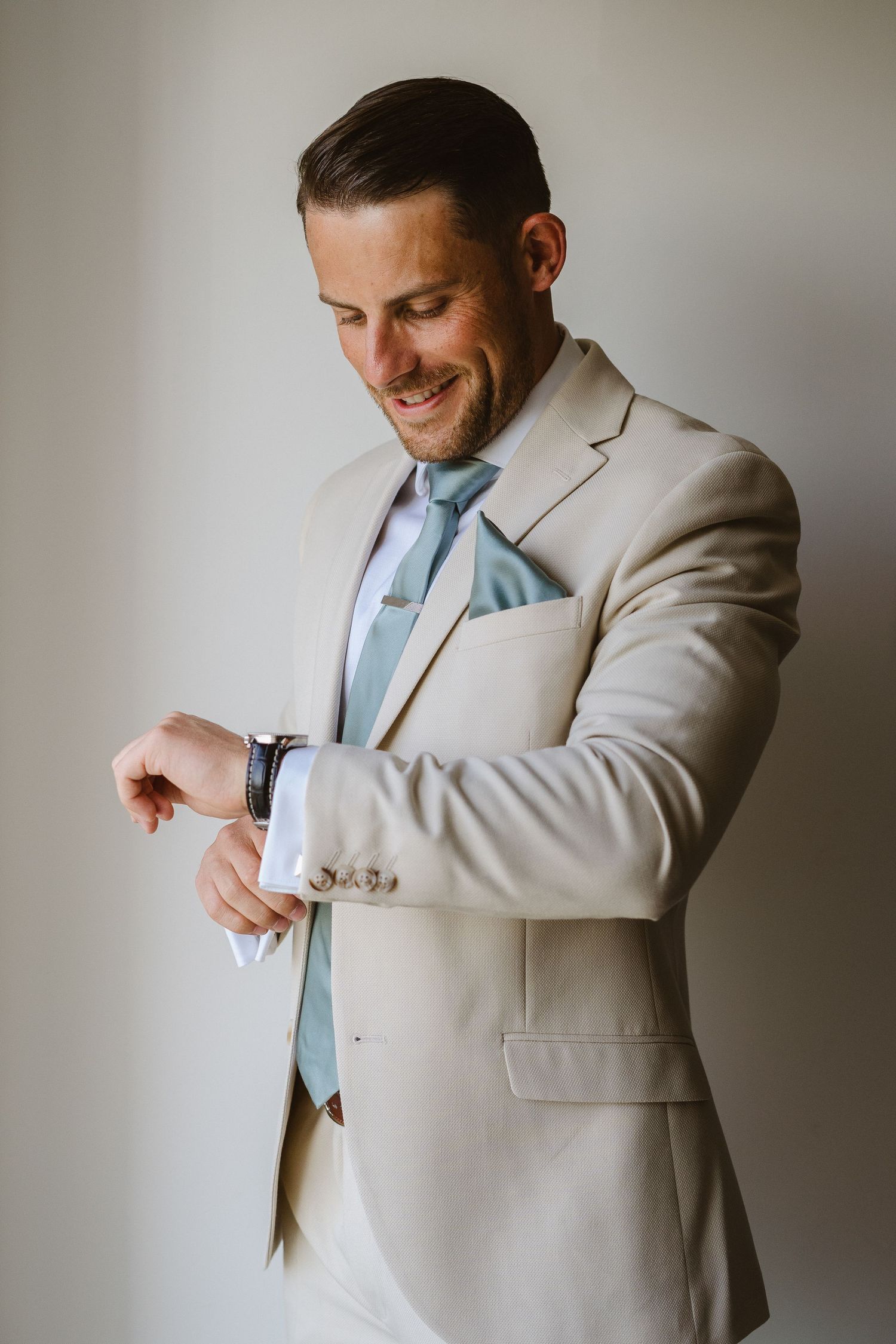 Groom in light beige suit and blue tie checks his watch while smiling during wedding preparations.