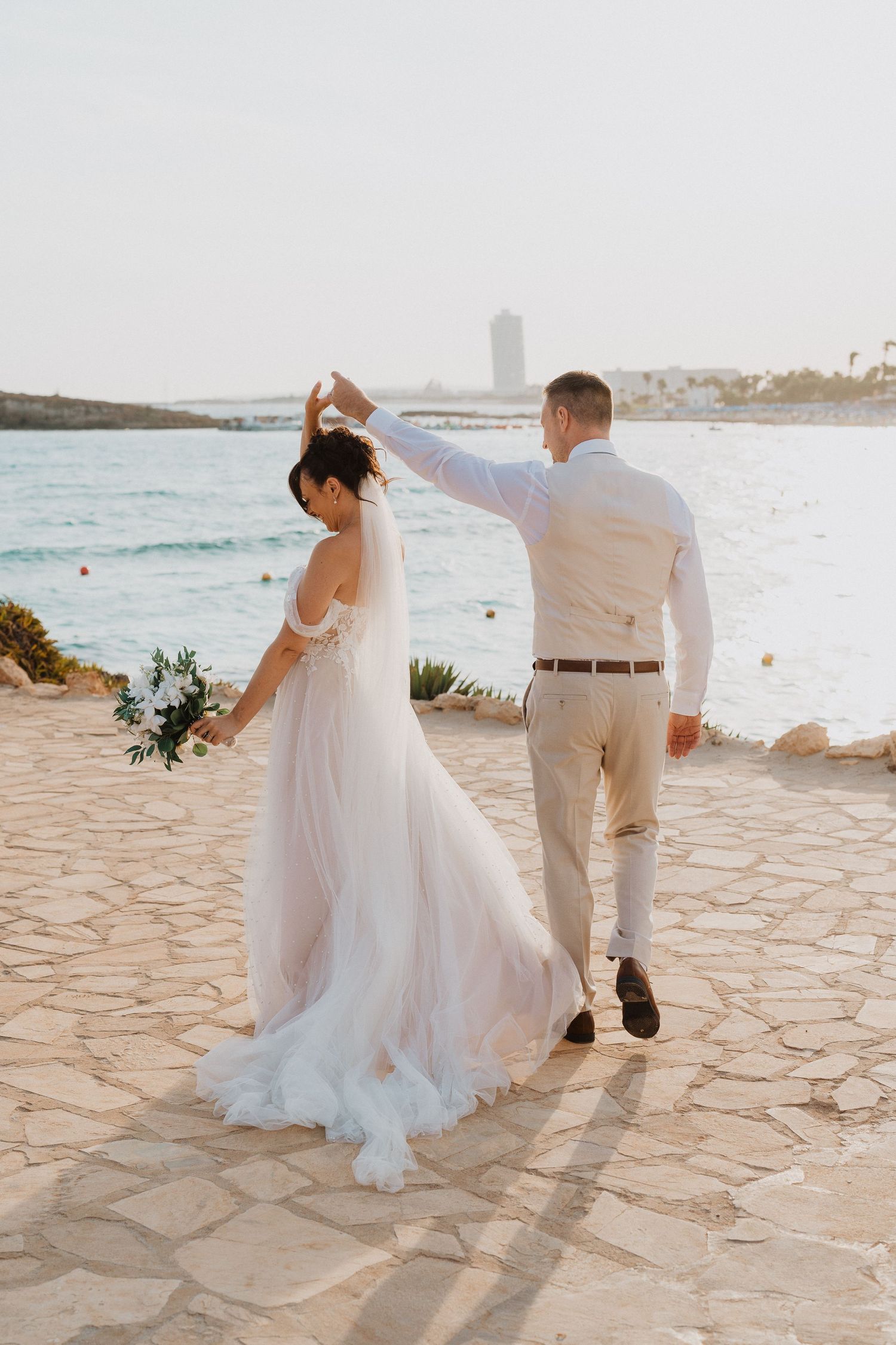 A couple in wedding attire dancing together on a coastal path with ocean and buildings in the background at sunset.