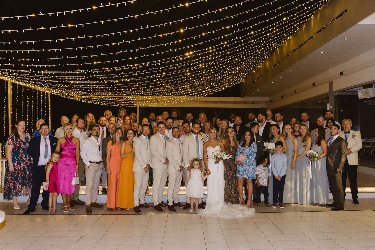 Wedding party and guests pose for group photo under romantic string light ceiling decoration.