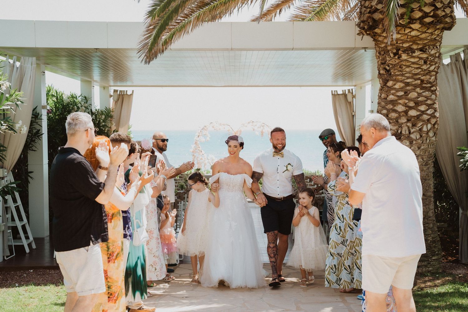 Wedding party walks through a palm-lined path as guests celebrate.