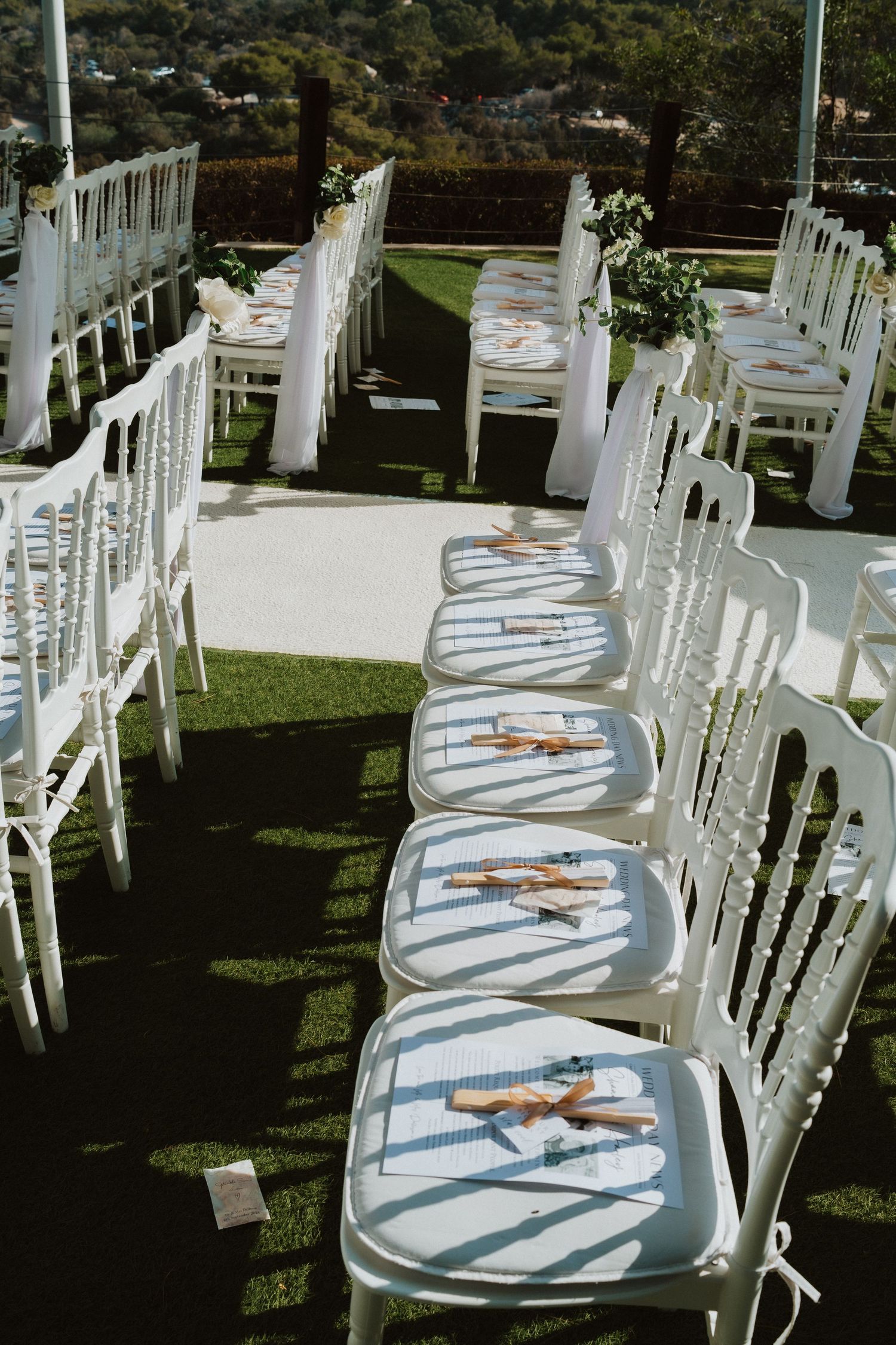 White ceremony chairs with floral decorations arranged in rows for an outdoor wedding.