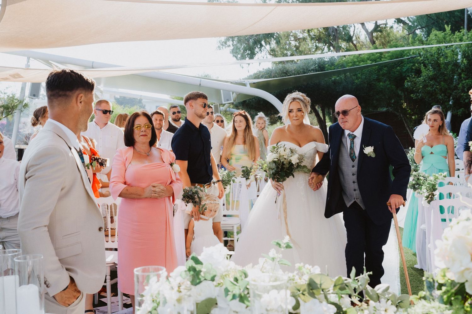 A wedding ceremony under a white canopy with elegant floral decorations and guests wearing formal attire.