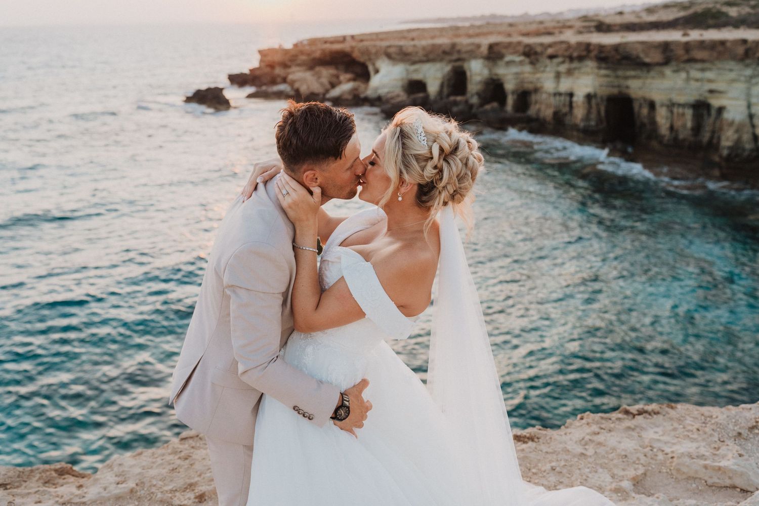 A romantic moment captured on a coastal cliff with turquoise waters and rocky shores in the background.