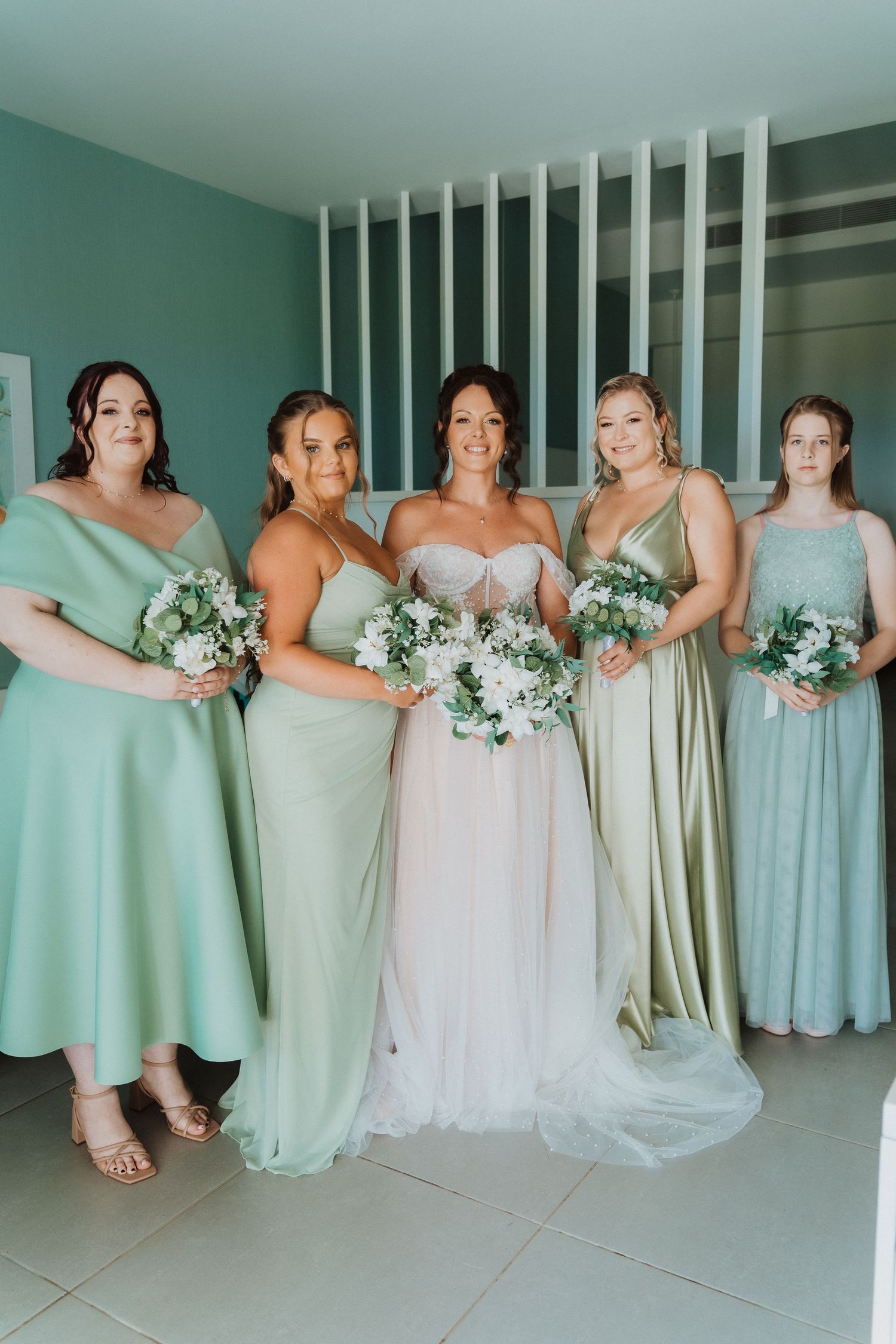 A bridal party wearing pastel mint and sage bridesmaid dresses poses together with matching bouquets.