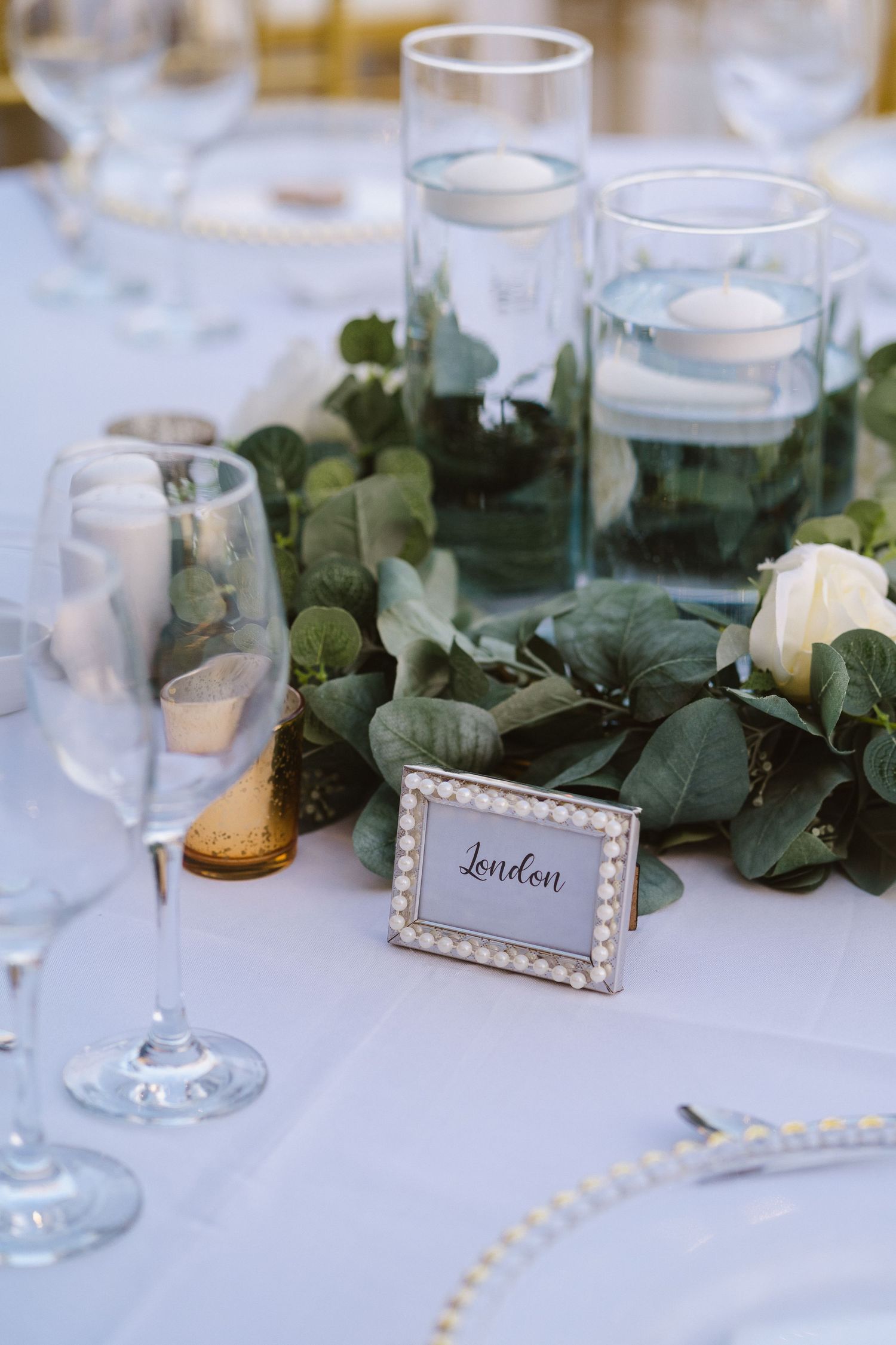 Pearl-framed 'London' table card set among greenery and glass votives at a formal dining setup.