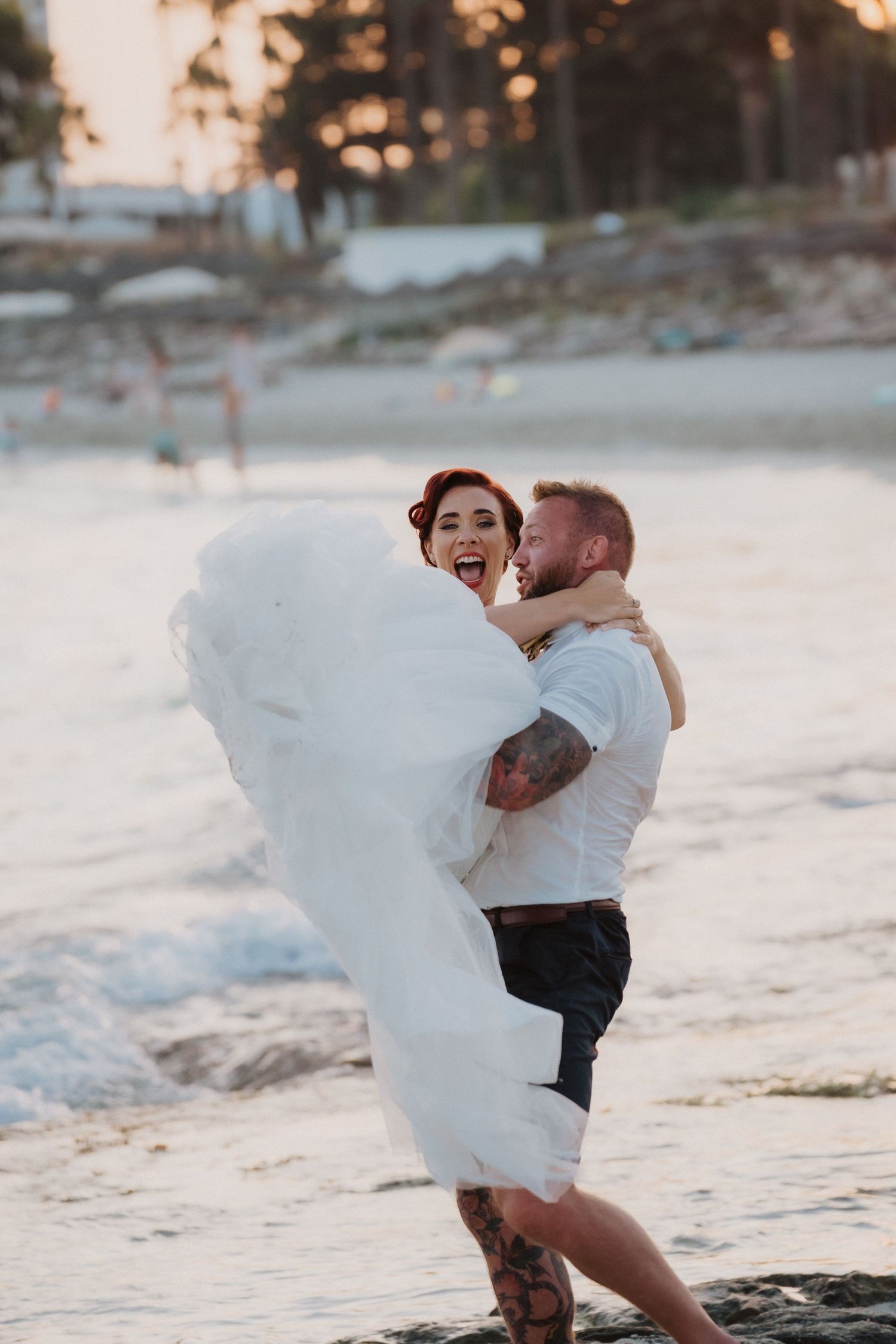 Joyful beach wedding moment with flowing white dress against sunset backdrop.