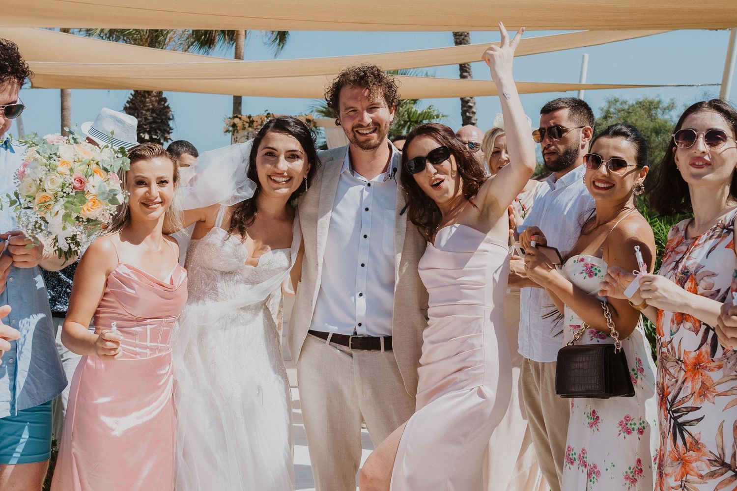 Wedding group poses for photo under beige canopy at beachfront venue.