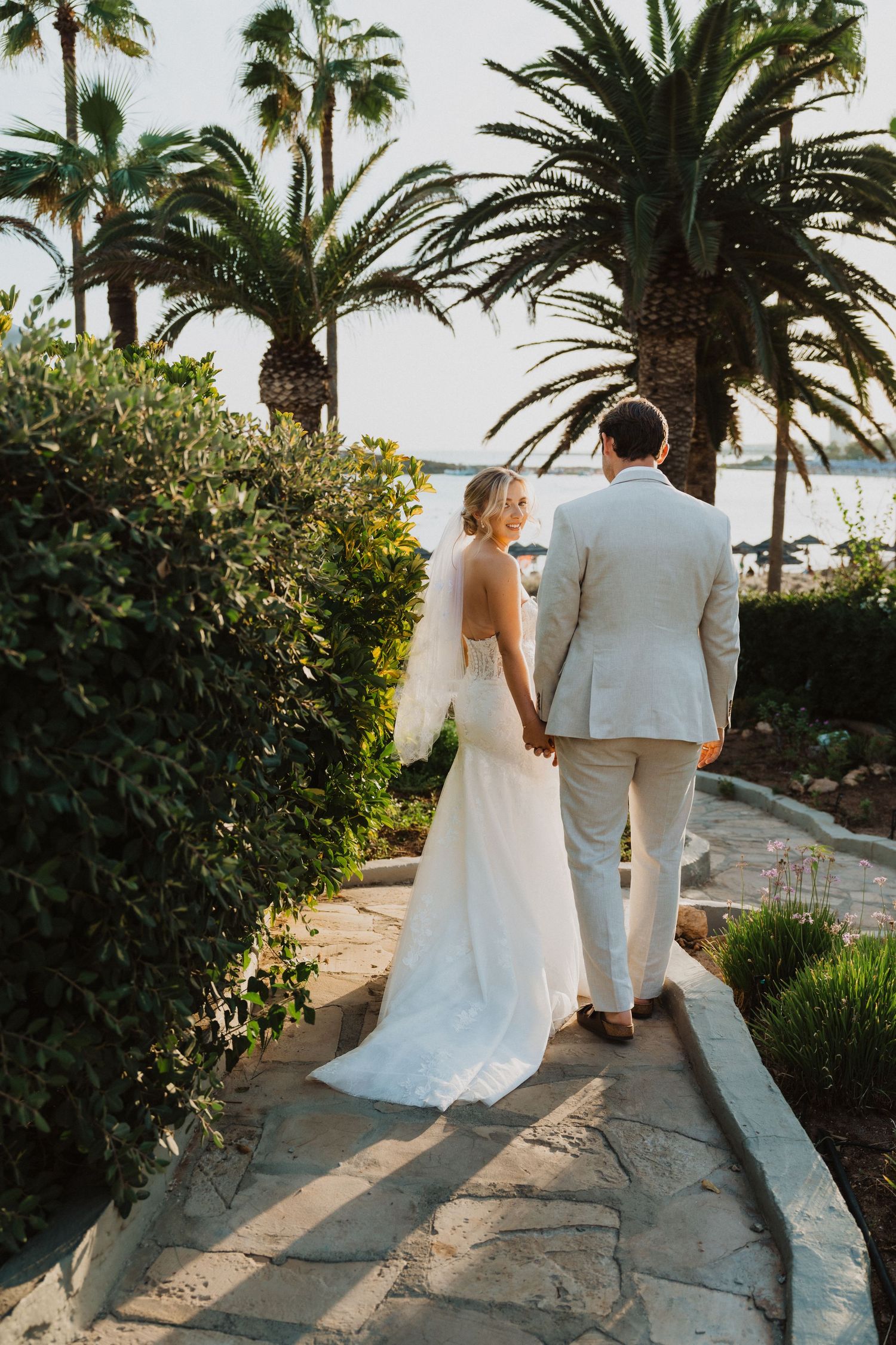 Bride and groom walking along coastal path with palm trees and ocean view.