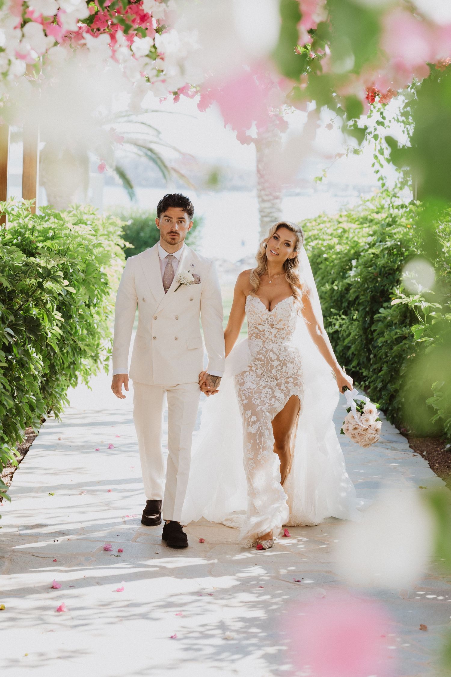 A bride and groom in white attire walk hand in hand along a sun-dappled path with pink flowers overhead.