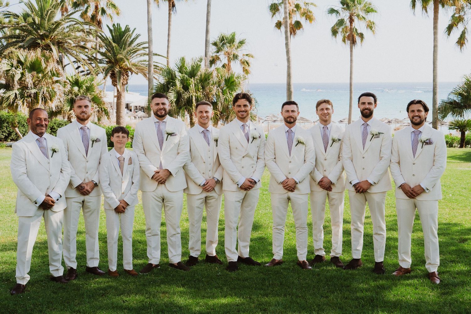 Wedding party in white suits poses on green lawn with palm trees in tropical destination setting.