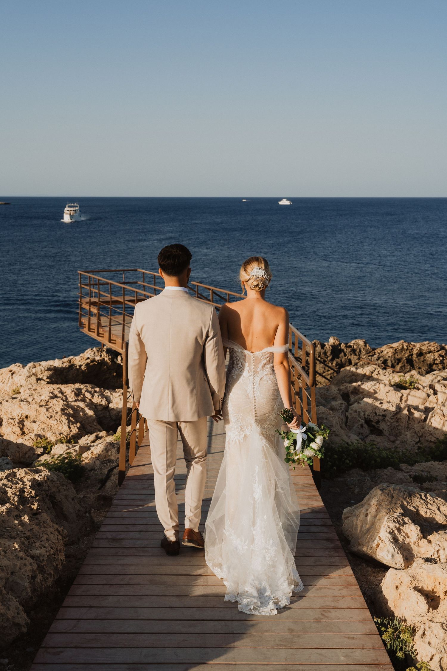 Couple walks along wooden boardwalk overlooking deep blue Mediterranean waters.
