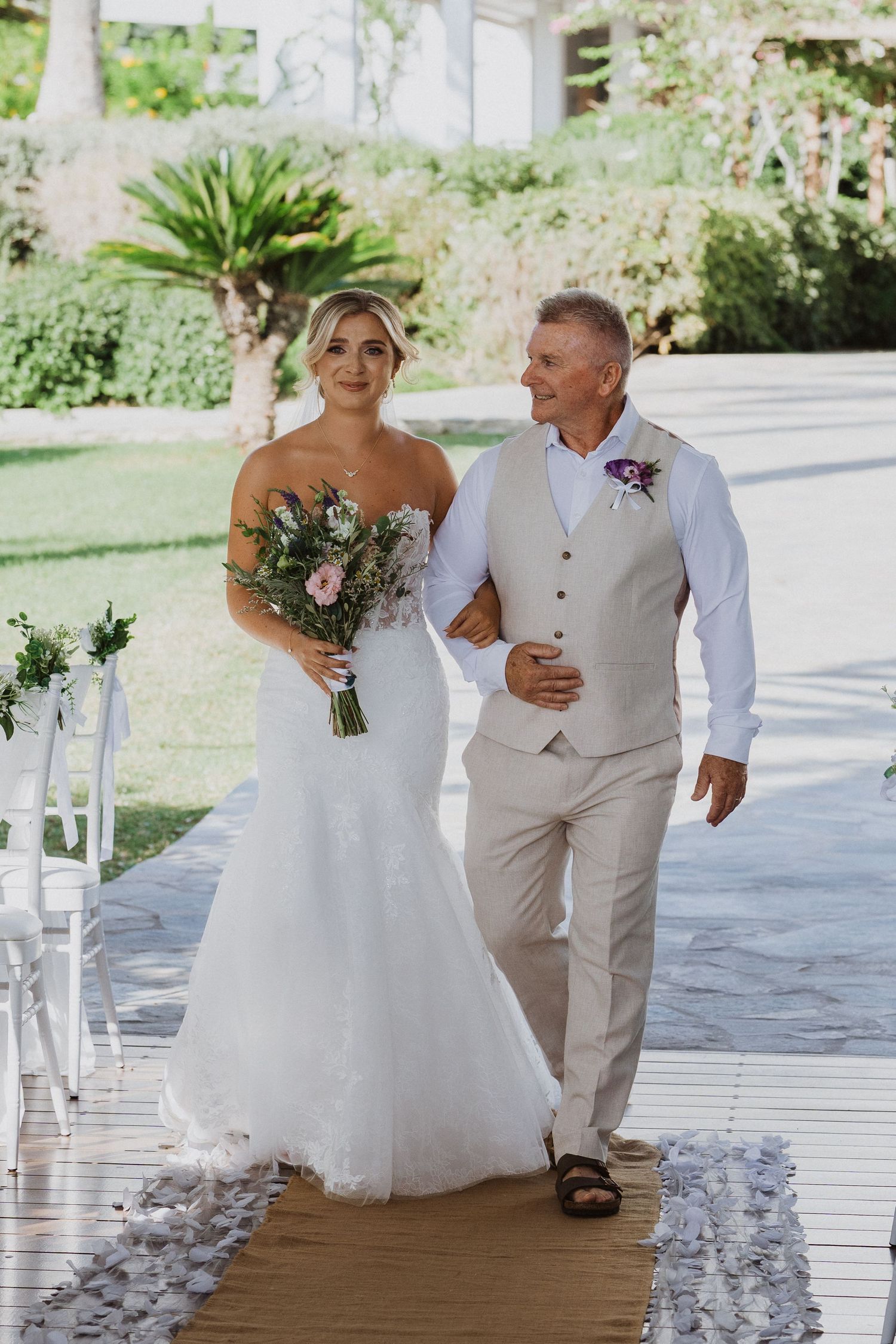 A wedding procession walks along a petal-lined aisle under a covered outdoor pavilion.
