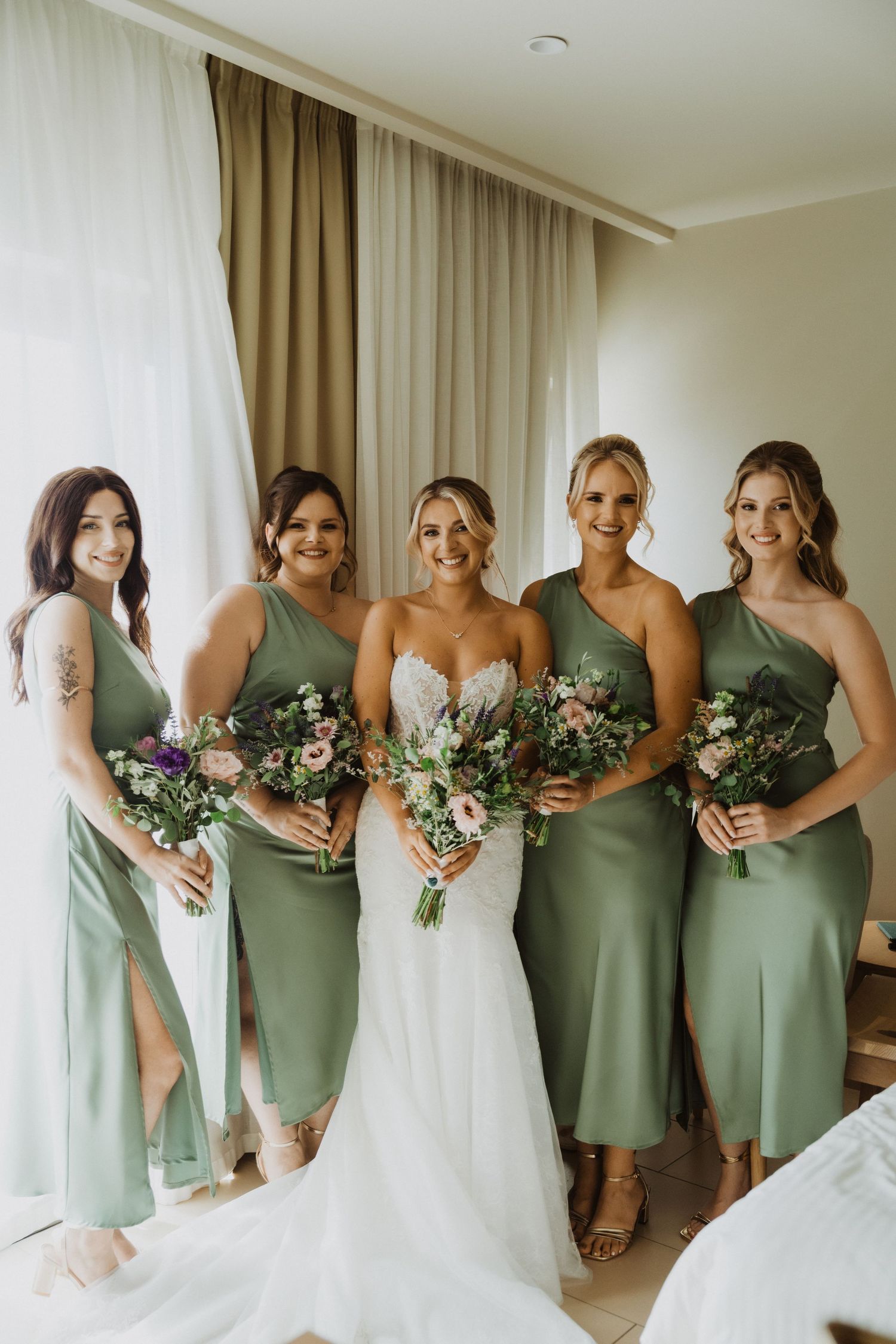 Joyful wedding party portrait with bridesmaids in sage green dresses and bride in white.