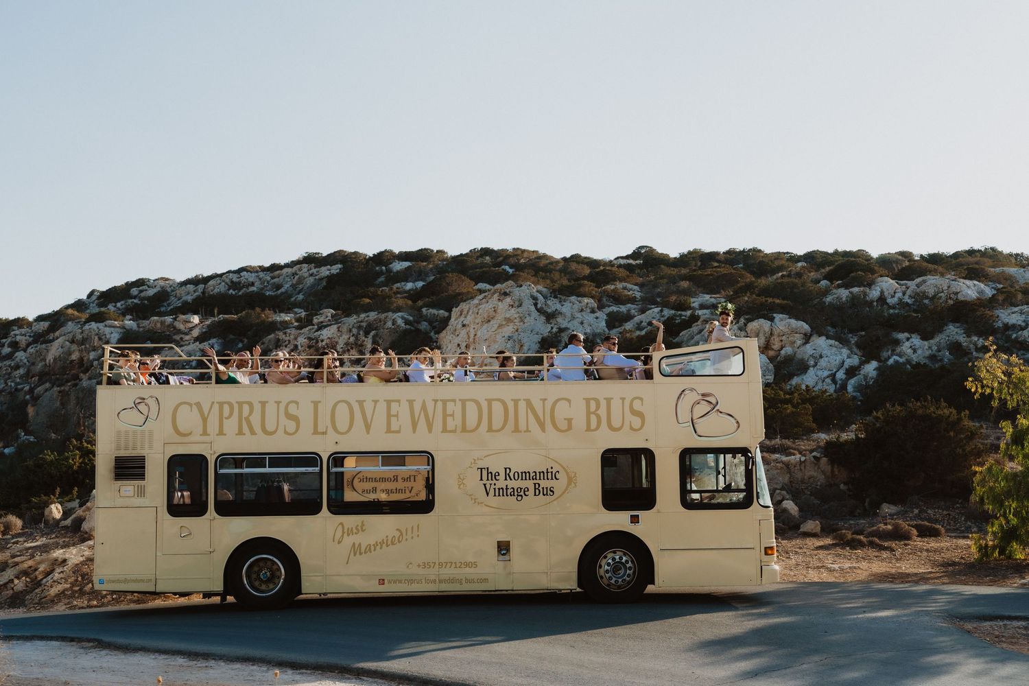 A long cream-colored wedding bus parked along a Mediterranean coastline.