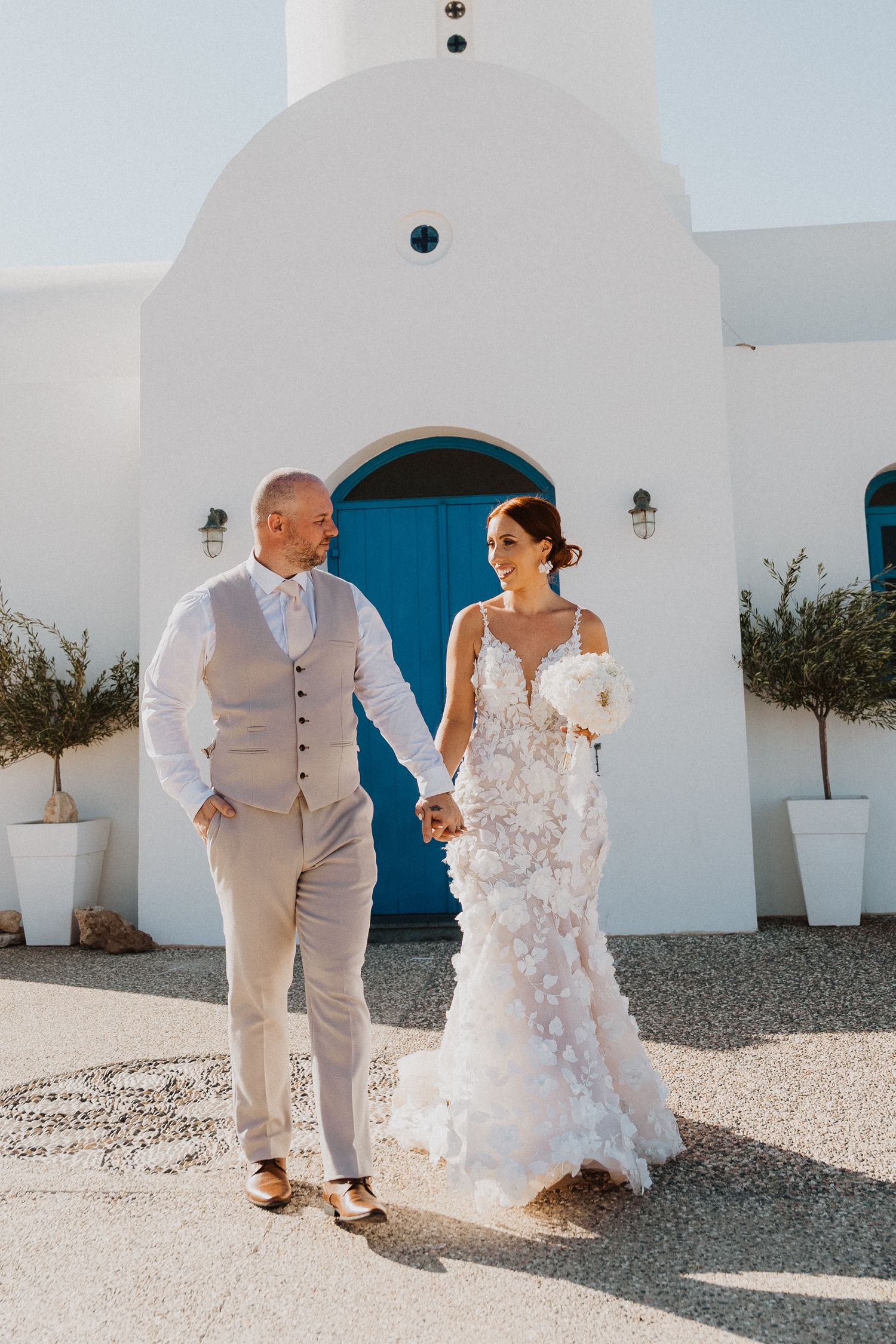 Newlyweds walking hand in hand outside a white Mediterranean church with bright blue door.