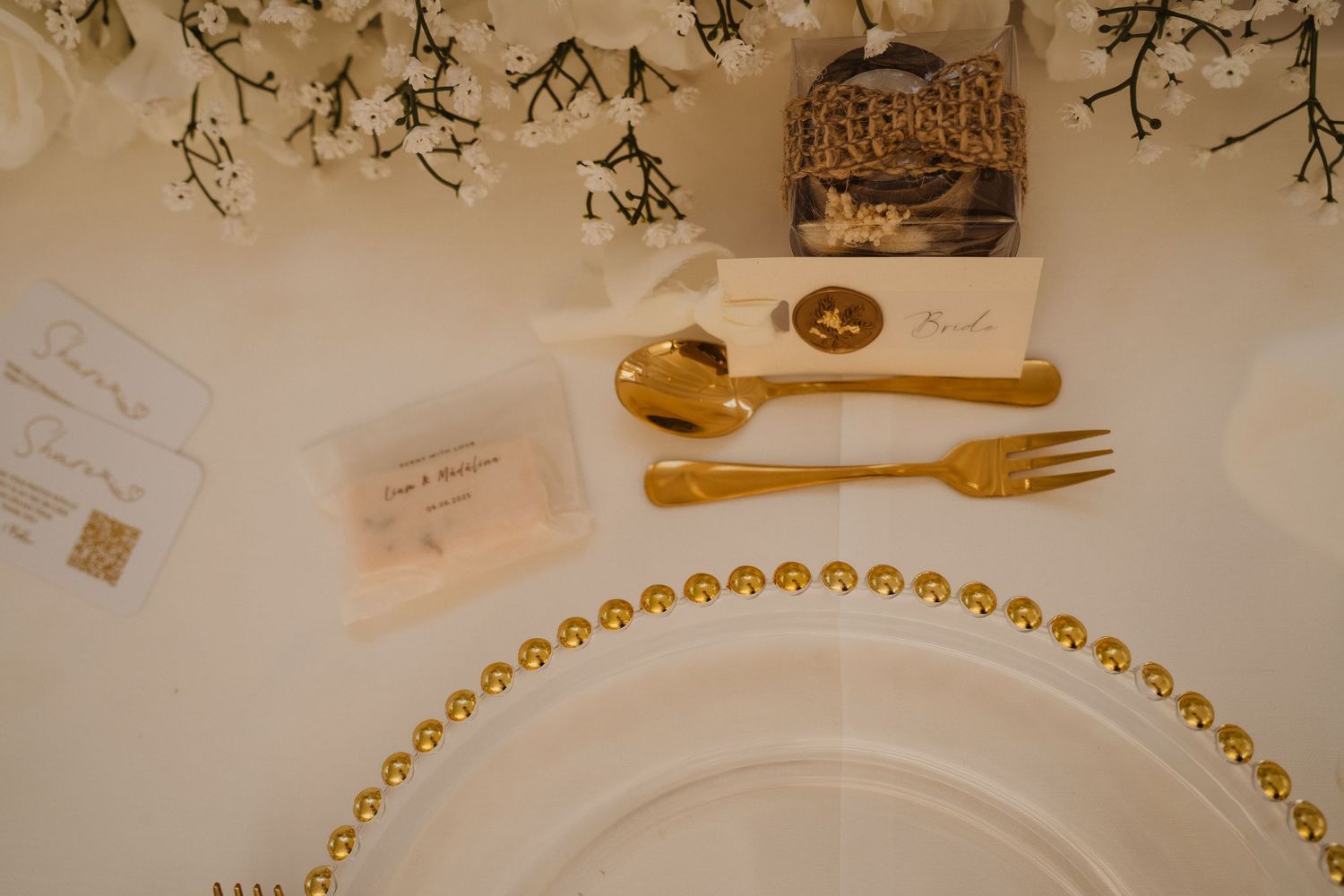 Elegant wedding table setting with gold cutlery, beaded charger plate, place cards, and floral decorations.