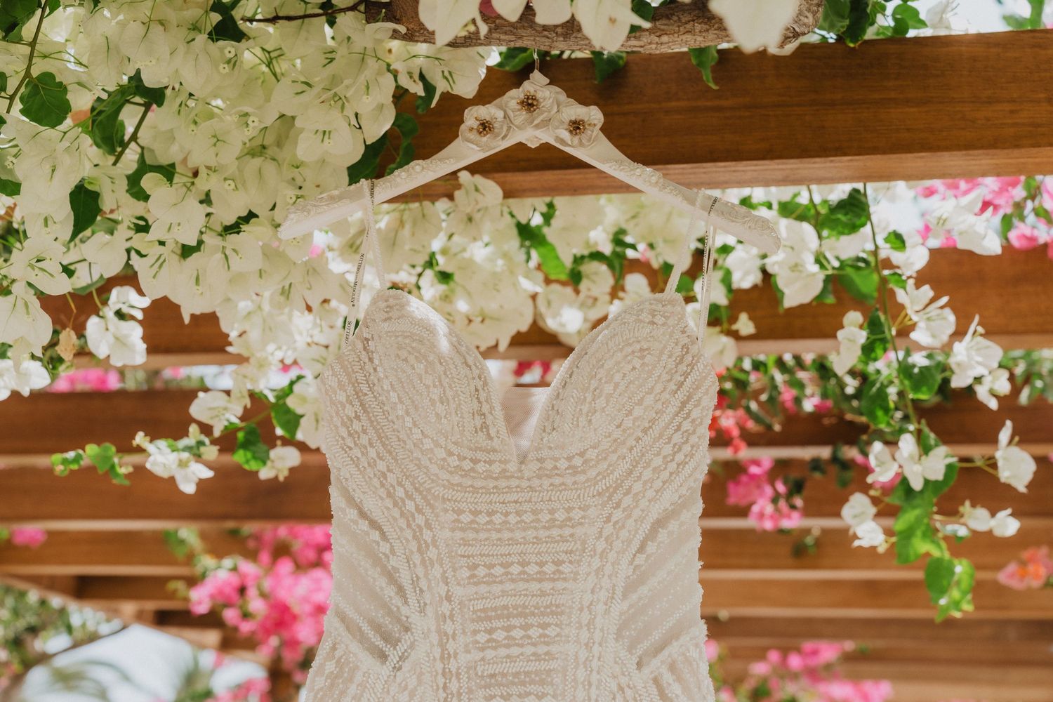 A lace wedding dress hanging on a wooden pergola adorned with white and pink bougainvillea flowers.