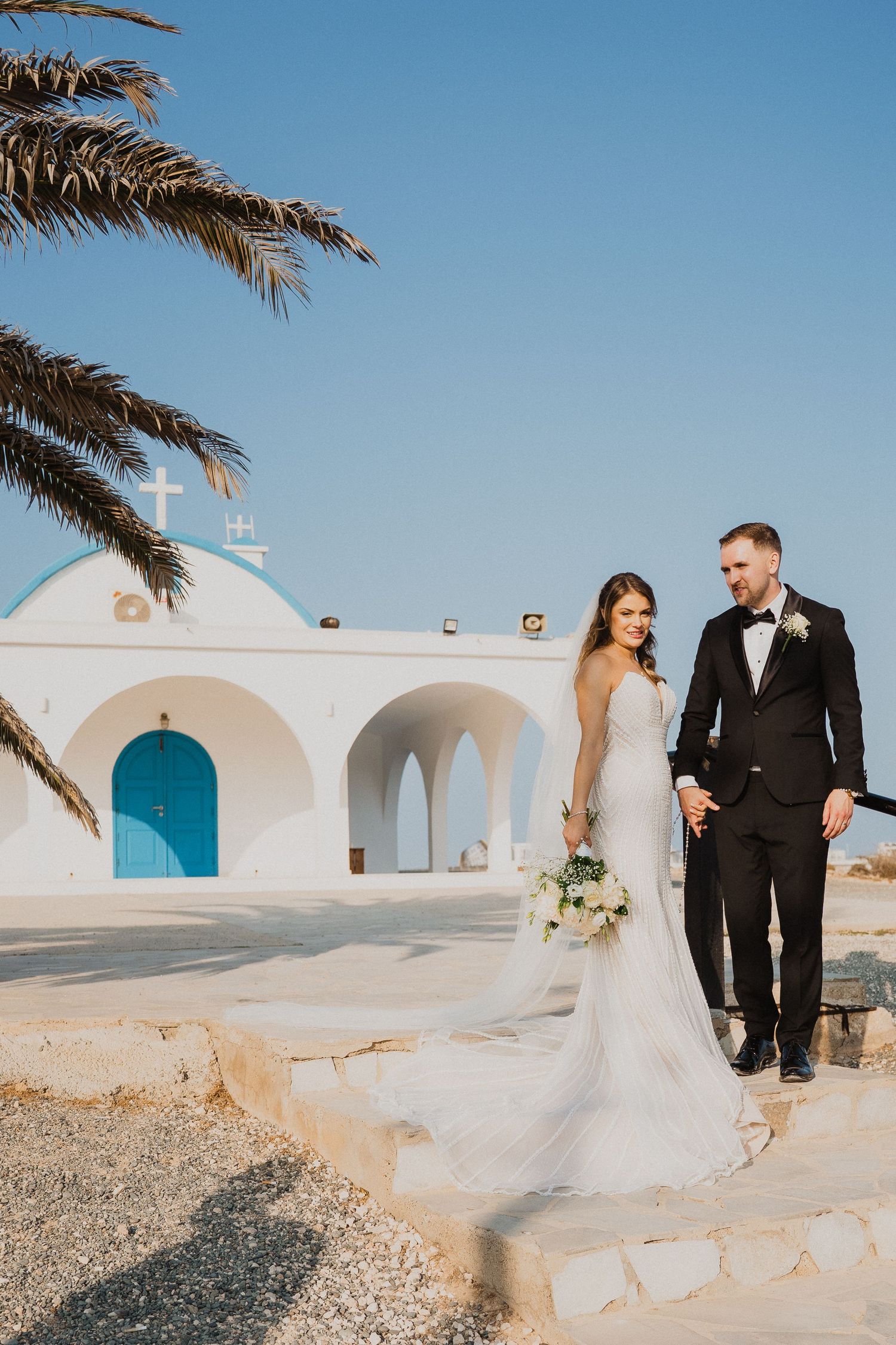 Bride and groom stand before a white Greek chapel with blue doors and a cross under clear blue sky.