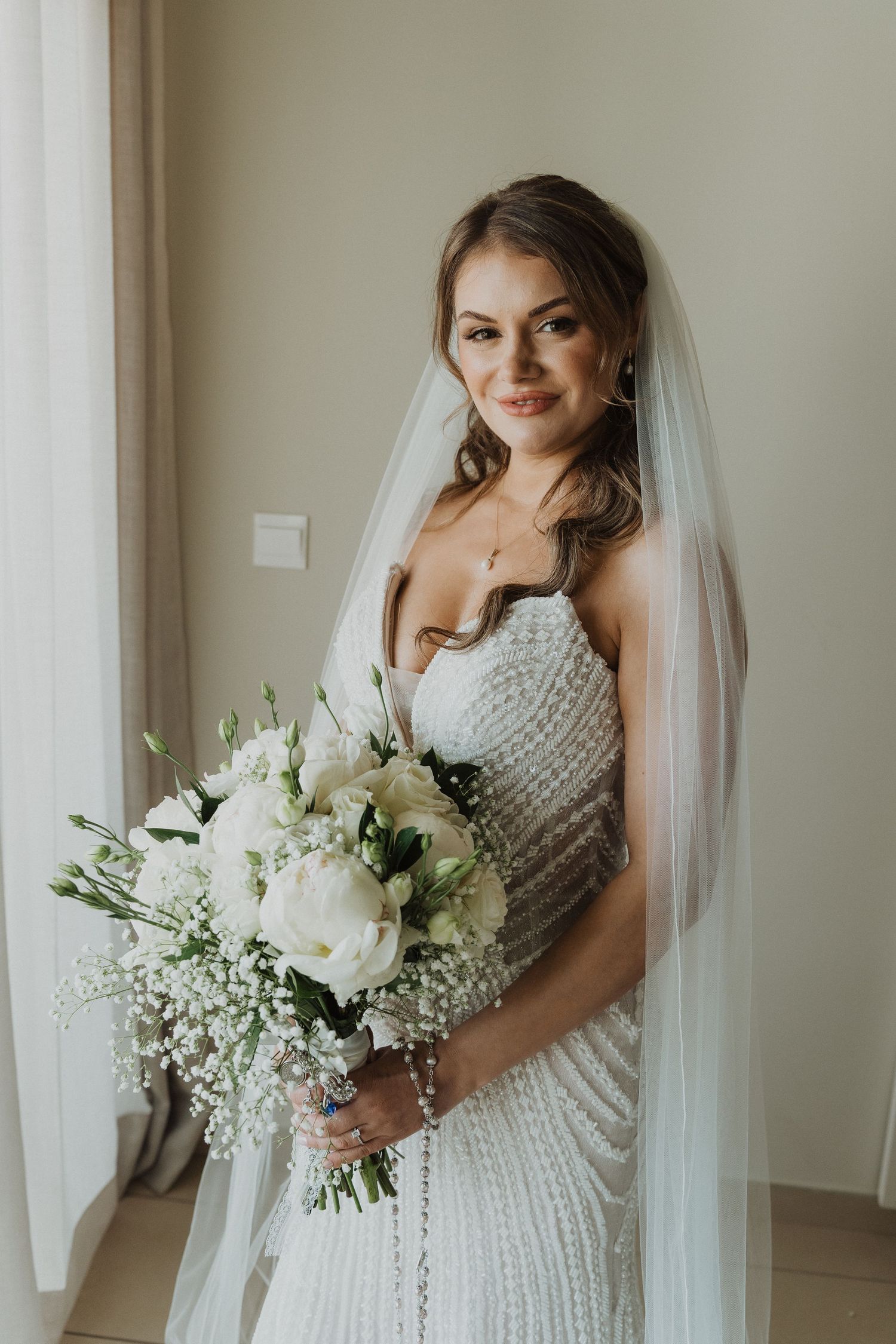 Elegant bride in beaded strapless gown and veil holding a lush white floral bouquet indoors.