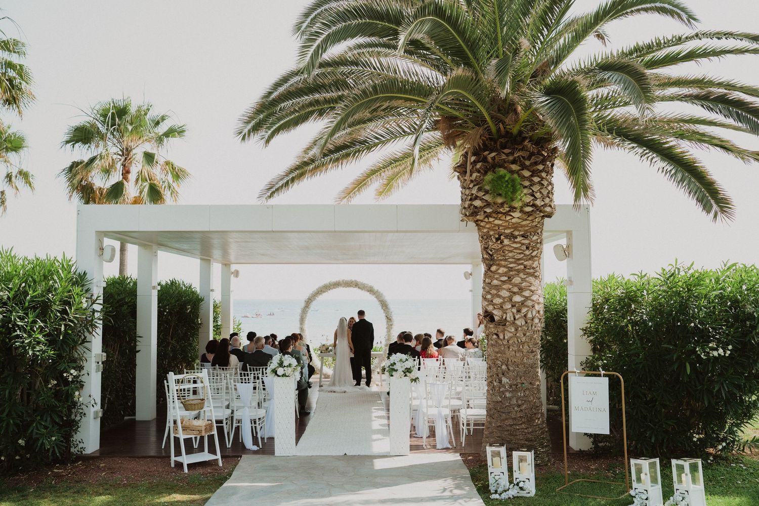 Wedding ceremony under white pergola with palm tree and ocean backdrop.
