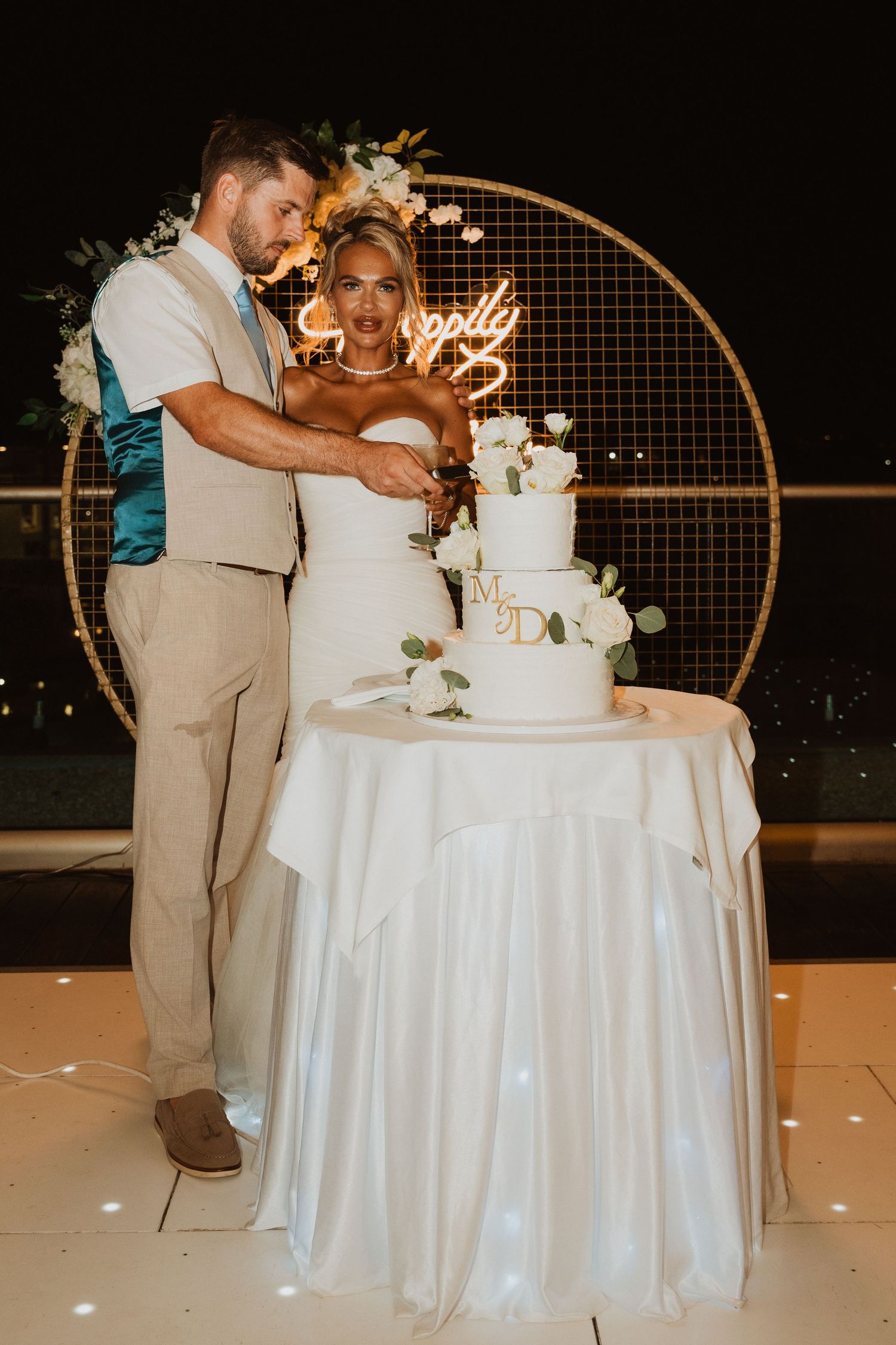 Bride and groom cutting their white wedding cake at an outdoor night reception with a glowing neon sign backdrop.
