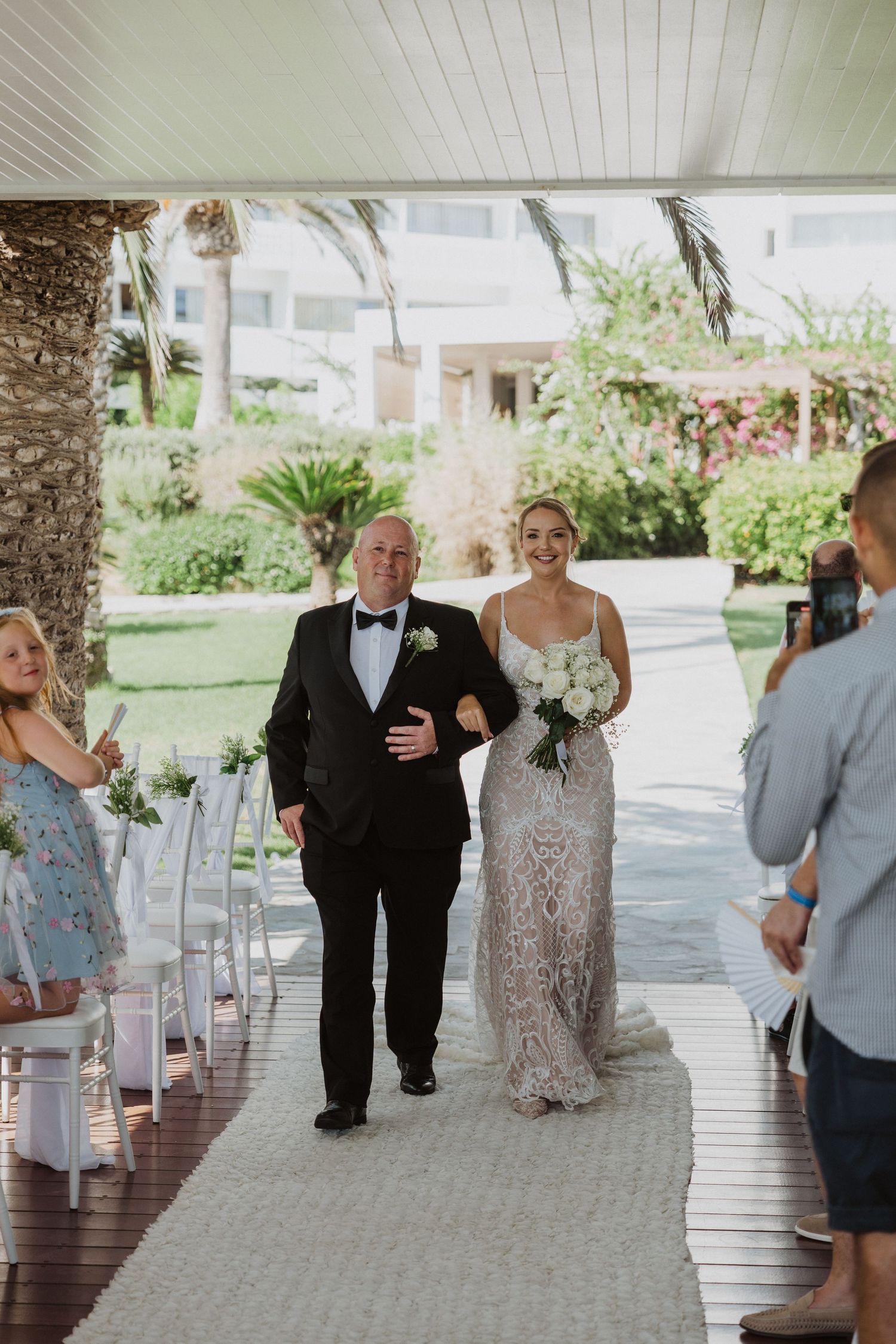 Wedding party members walk down a covered outdoor pathway lined with white chairs and tropical landscaping.