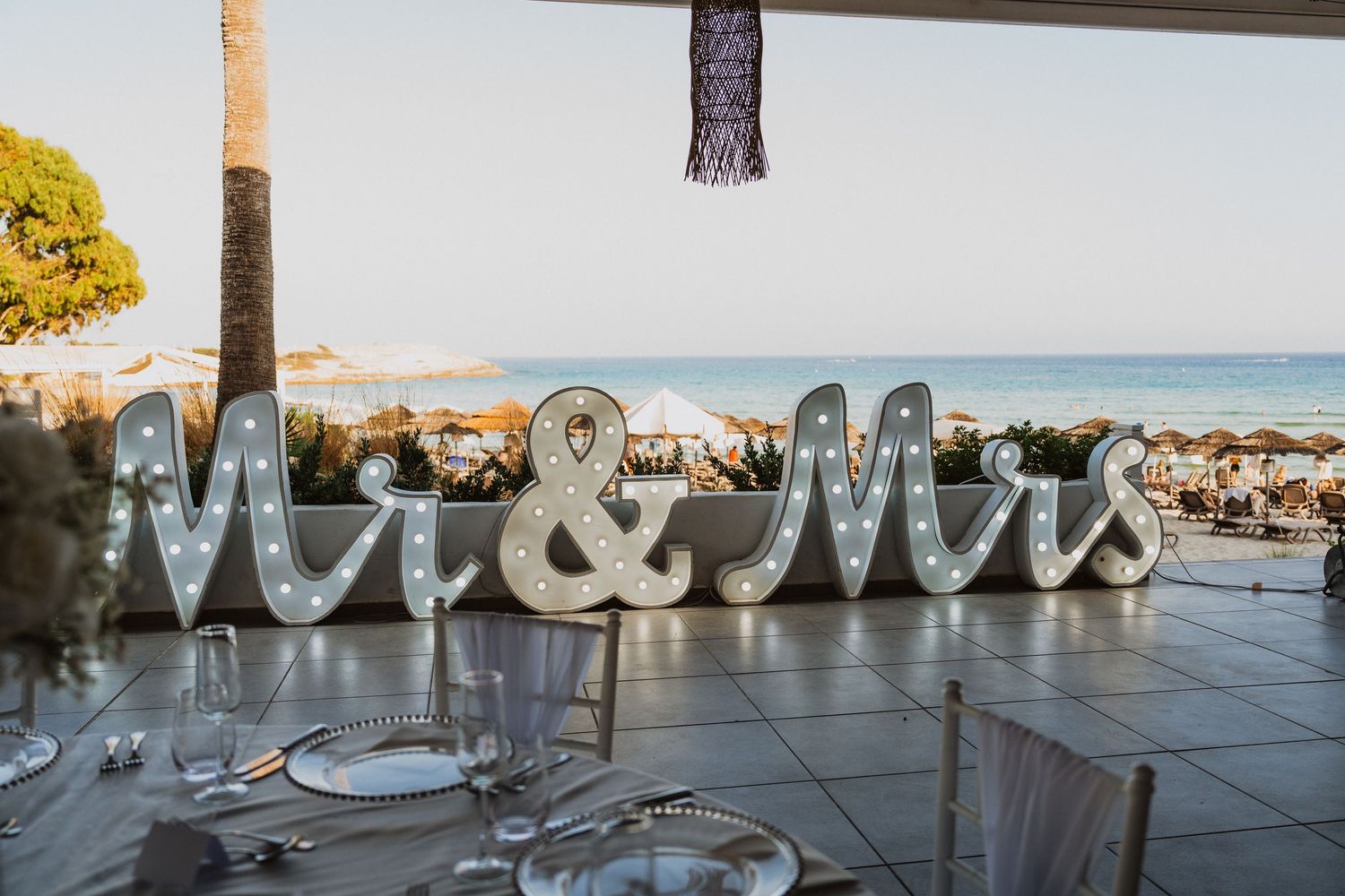 Illuminated 'Mr & Mrs' marquee letters on display at an outdoor wedding reception overlooking the ocean at sunset.