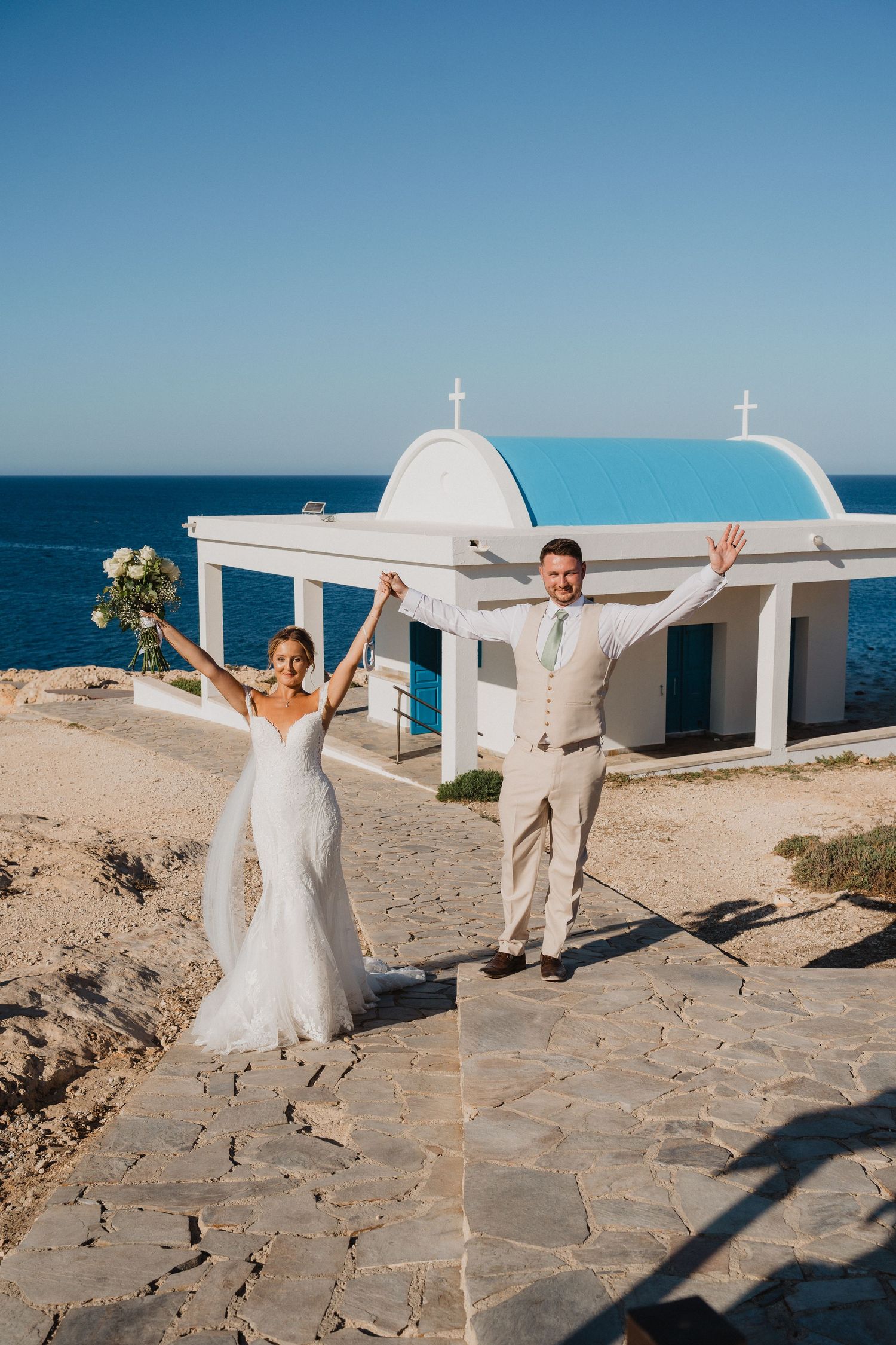 A wedding couple poses joyfully in front of a white Mediterranean chapel by the ocean on a sunny day.